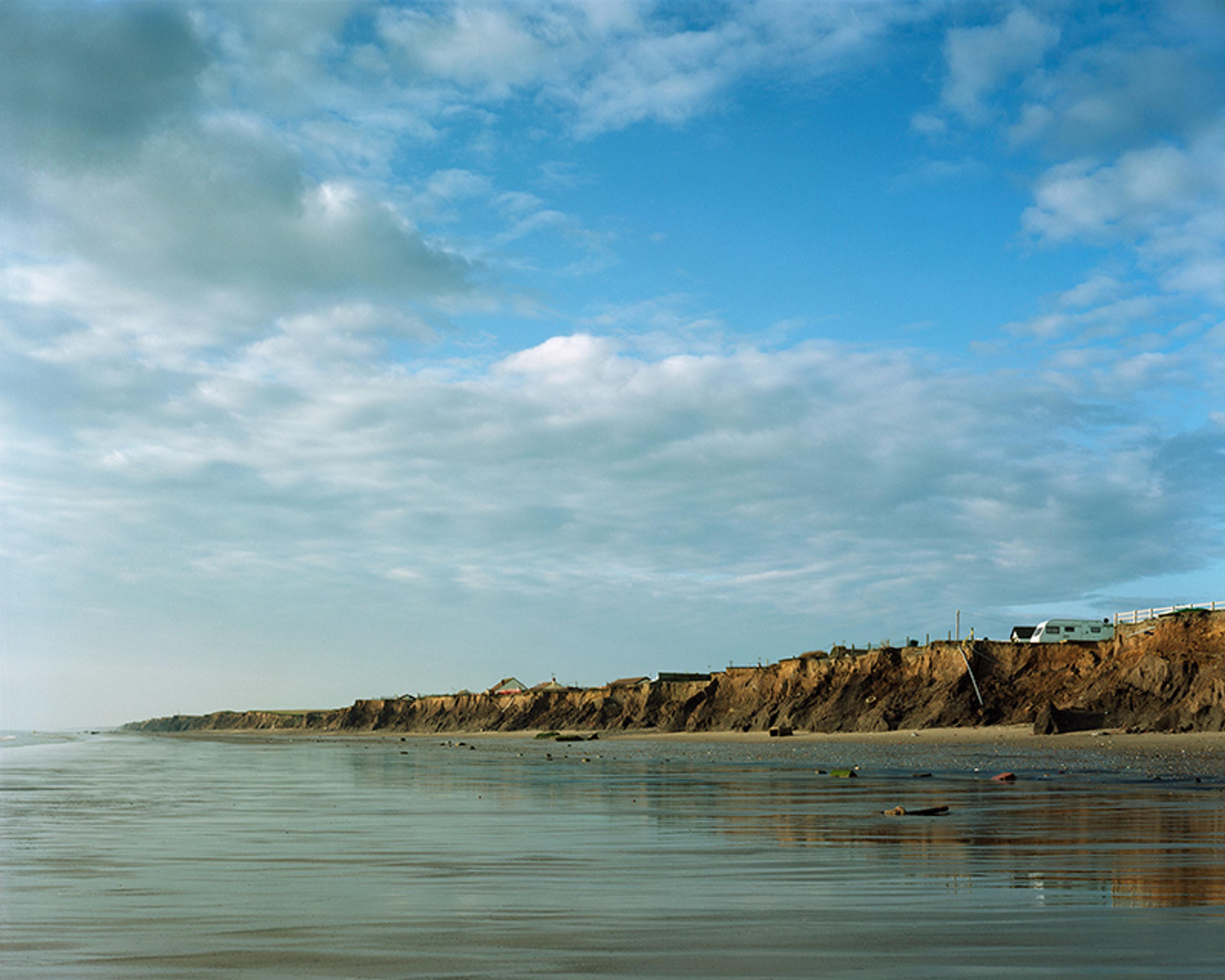 A sandy beach with eroded cliffs under a blue sky. A campervan is parked atop the cliffs.