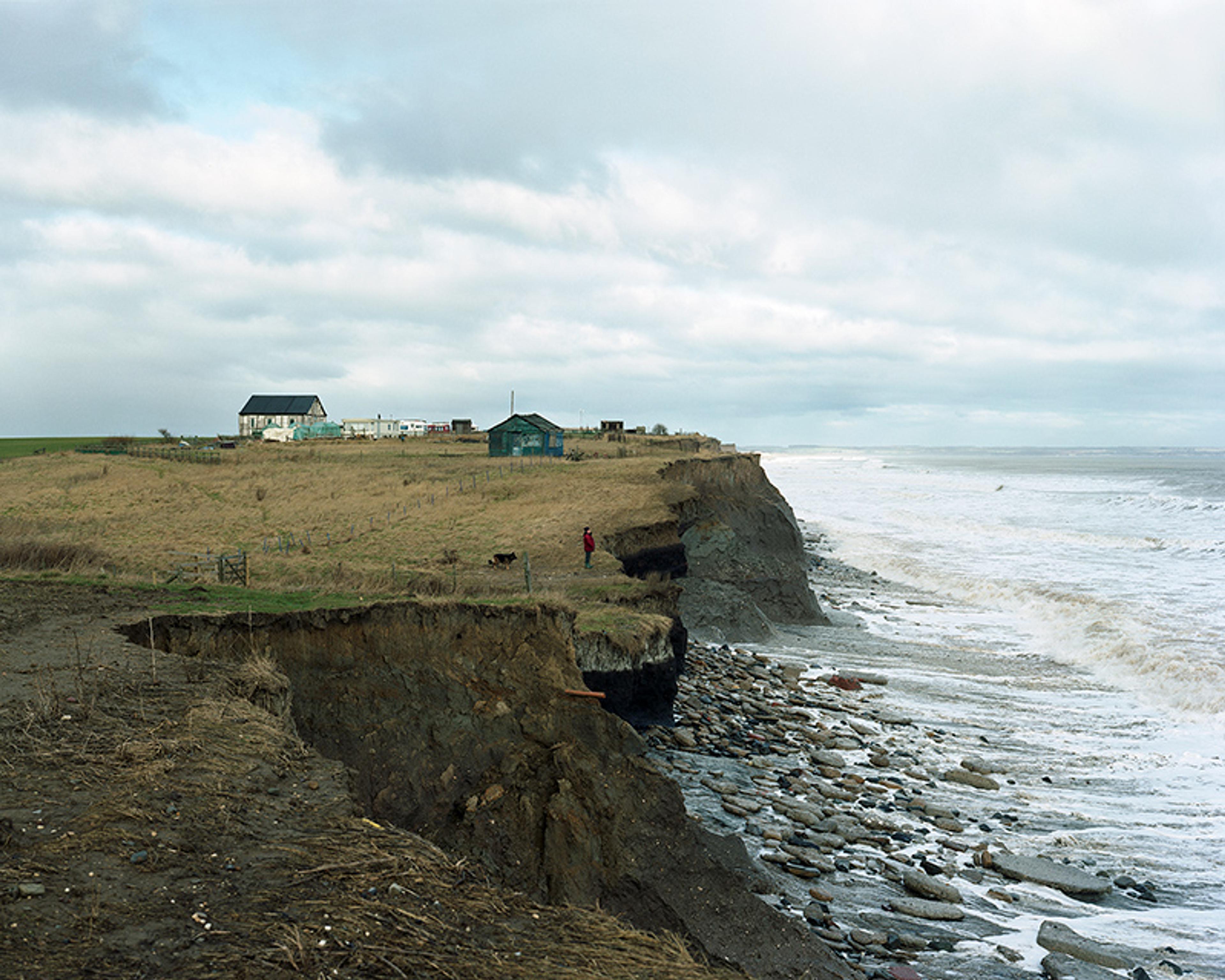 A coastal cliff with eroded edges, a person in red, houses on a grassy plateau, and a view of the ocean.