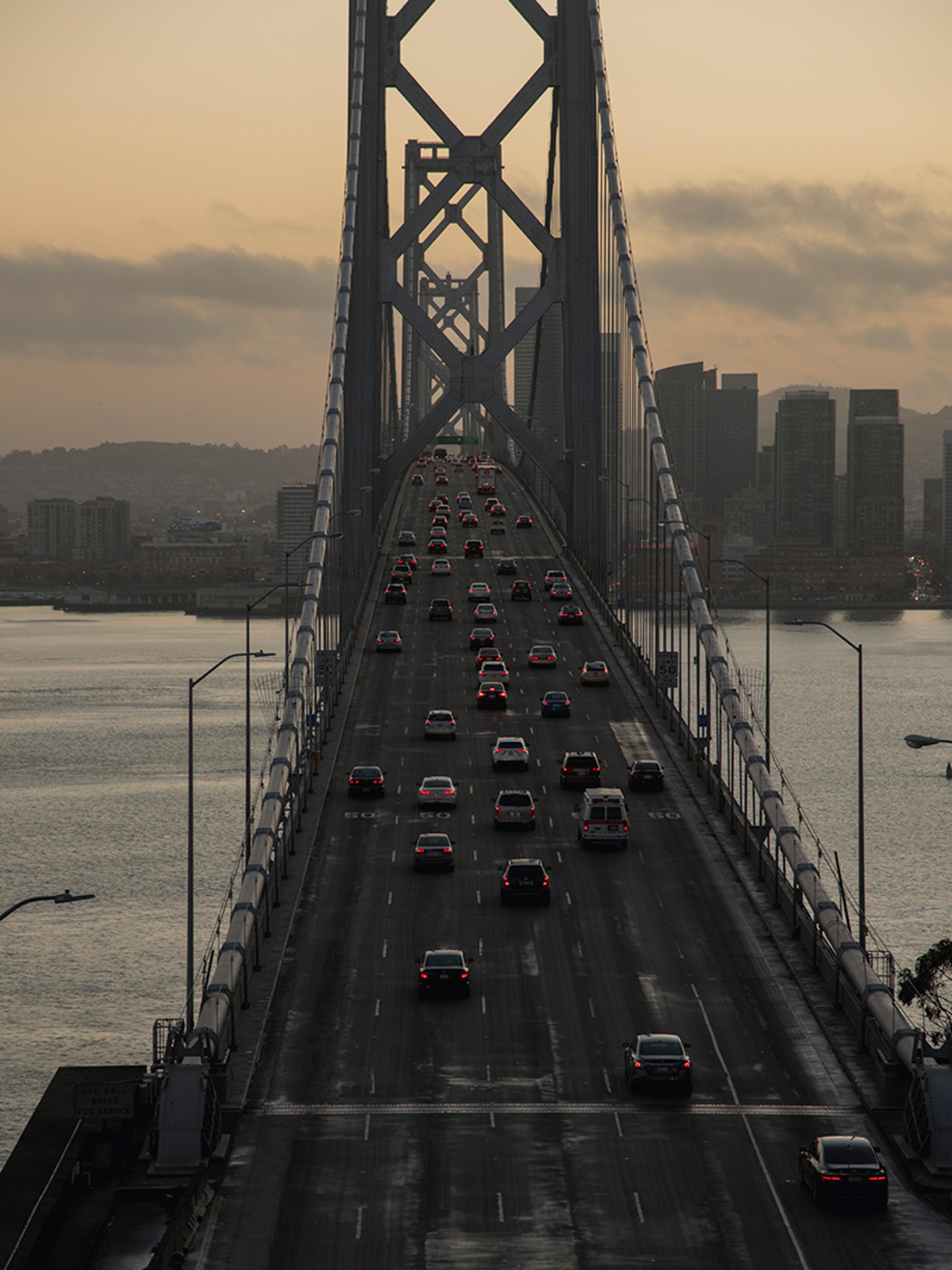 A busy bridge at dusk with cars, a city skyline in the background and a tranquil water view.