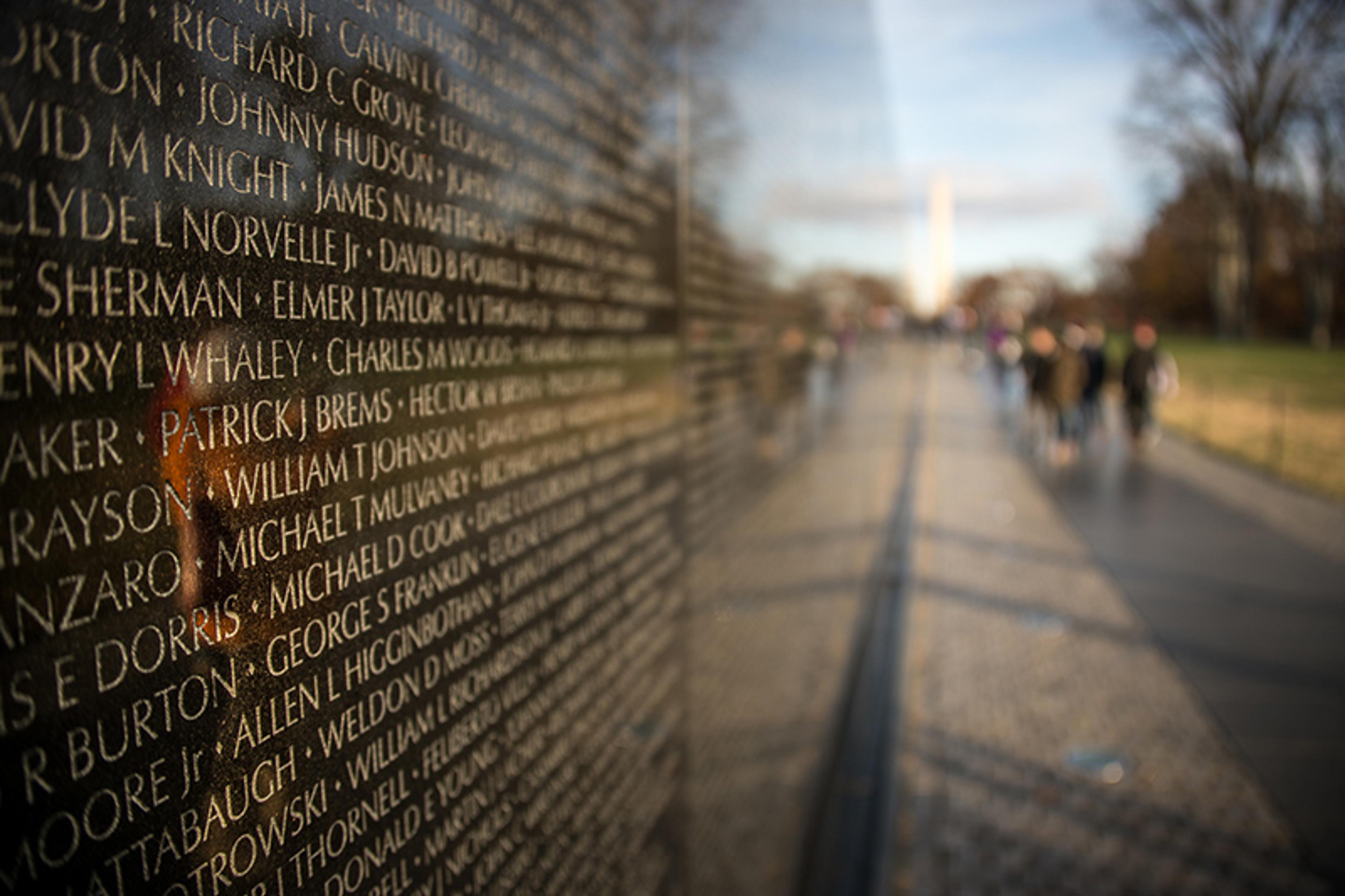 A reflective memorial wall with engraved names, blurred figures in the background, taken on a sunny day.