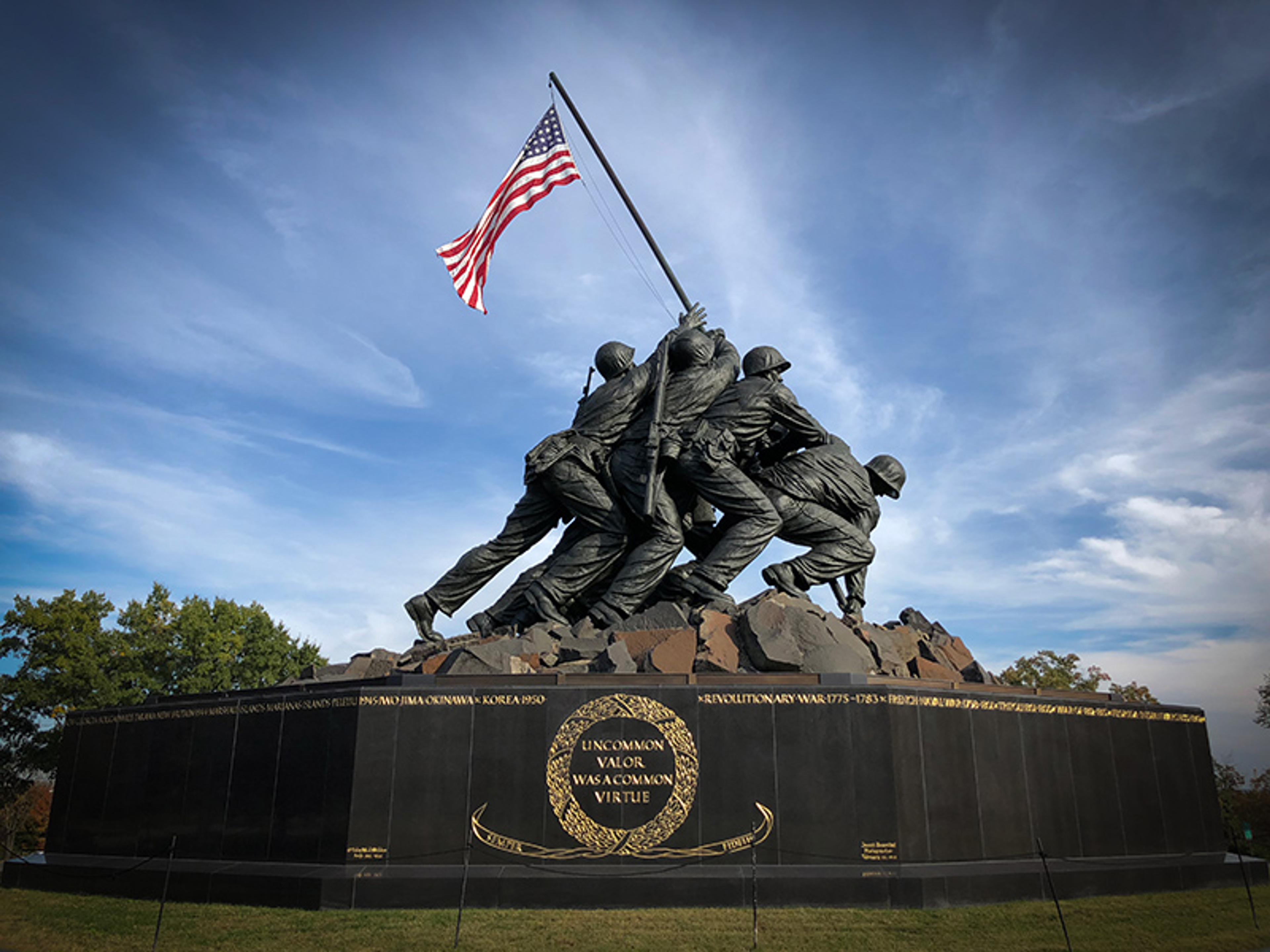 A bronze statue depicting soldiers raising a flag, set against a blue sky. The base has a gold-inscribed message, which reads uncommon valor was a common virtue.