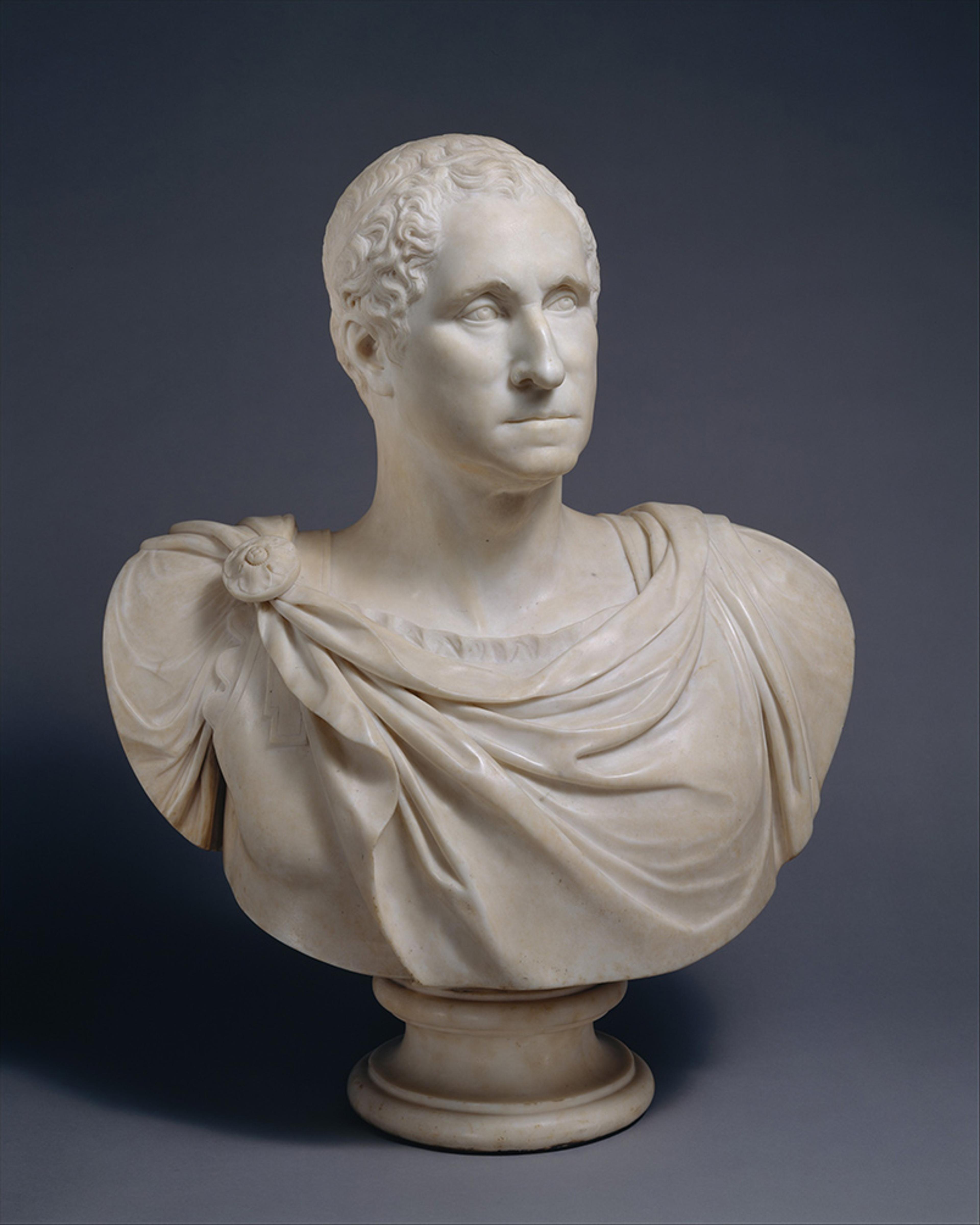 Marble bust sculpture of a man with curly hair wearing draped clothing on a pedestal against a dark background.