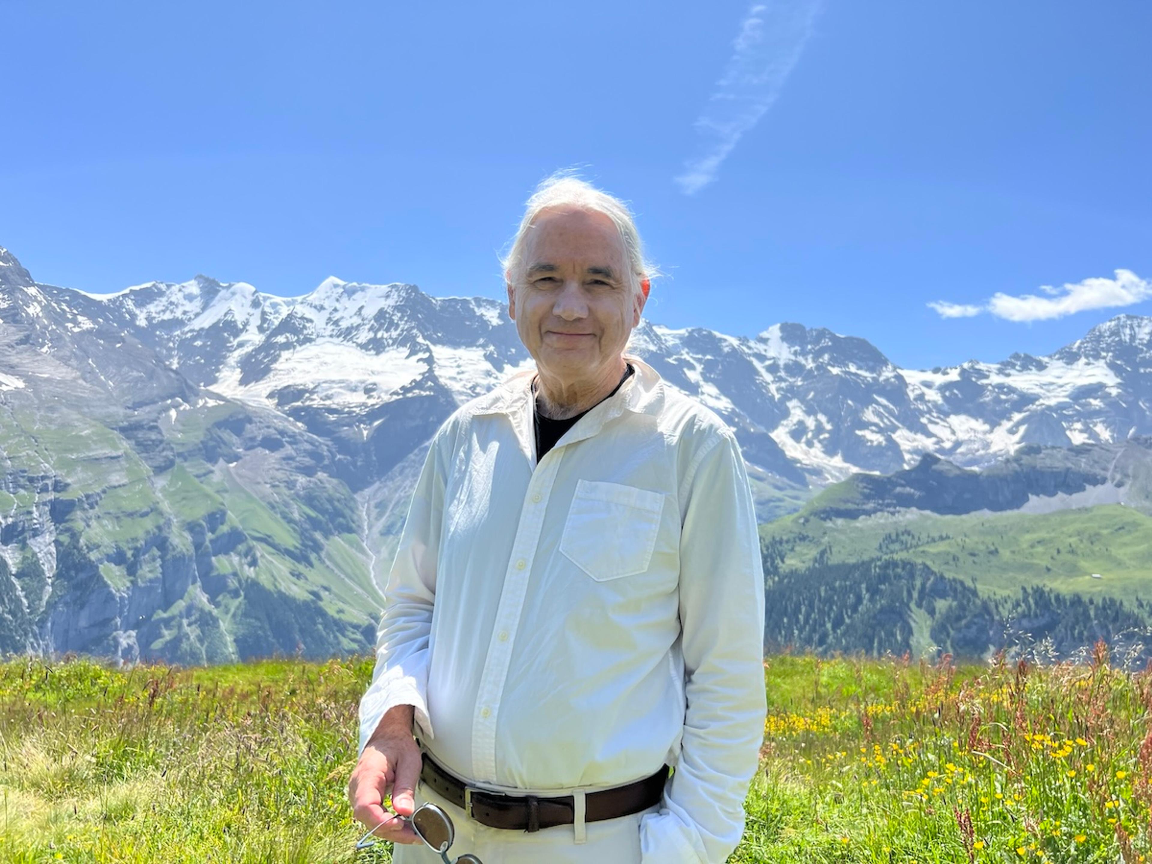 Photo of a man in a white shirt standing in a meadow with snow-capped mountains in the background under a clear blue sky.