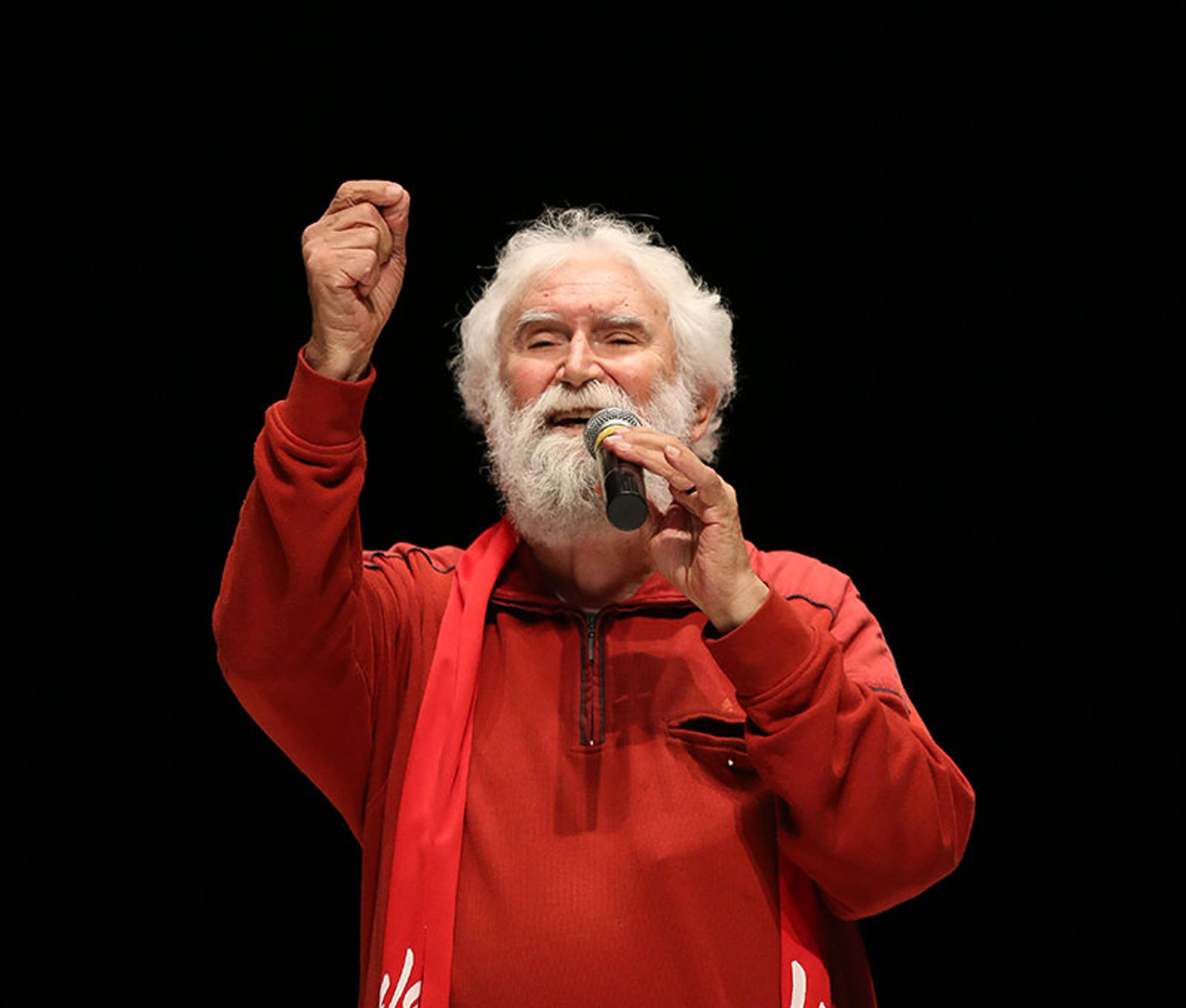 An older man with white hair and beard speaking into a microphone wearing a red jacket against a dark background.