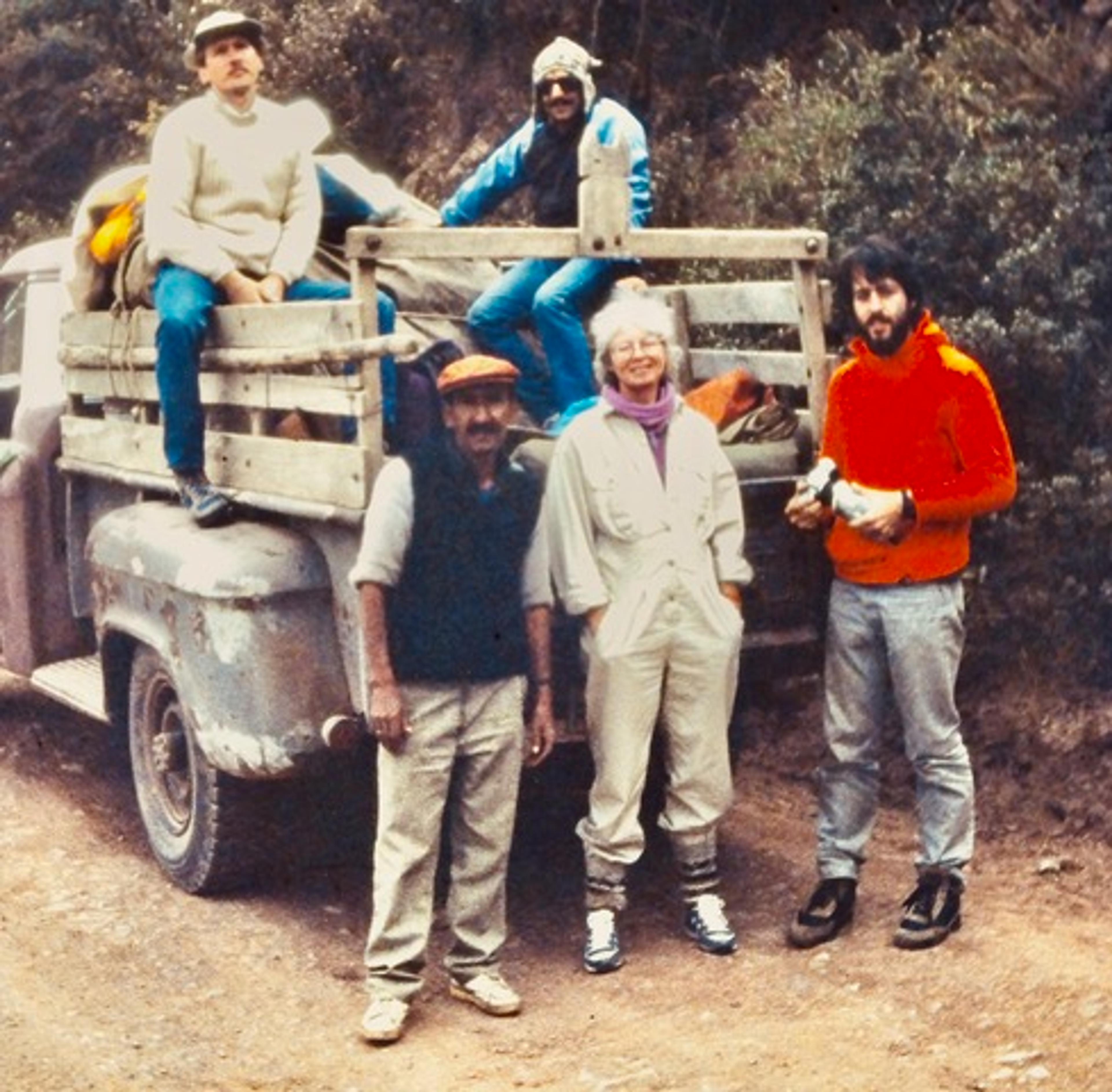 Photo of five people outdoors by a vintage truck, two seated on its bed. They are dressed for hiking or outdoor activity.