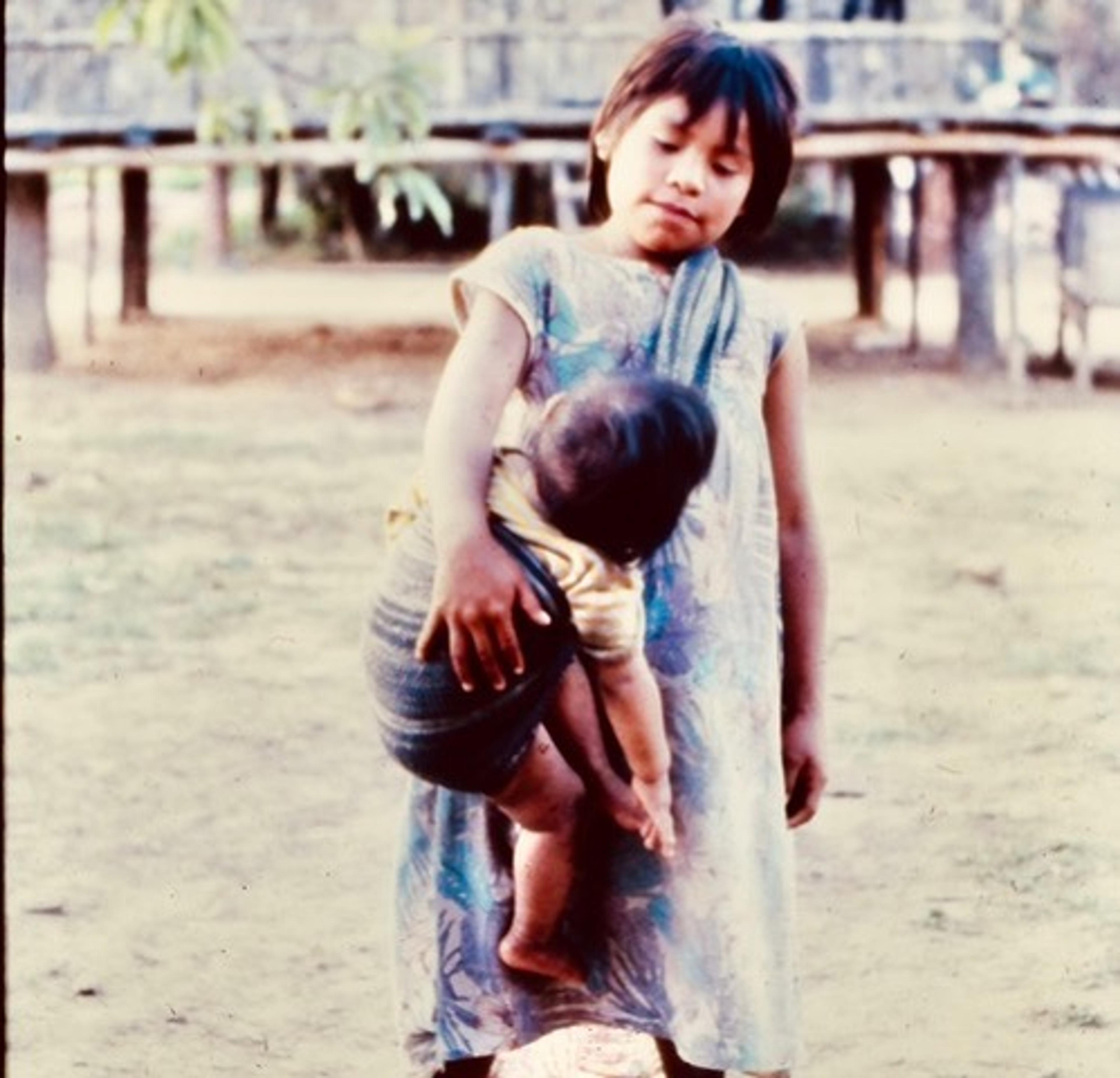 Foto de uma criança carregando um bebê em um sling, em pé ao ar livre em um caminho de terra com árvores e uma estrutura de madeira ao fundo.