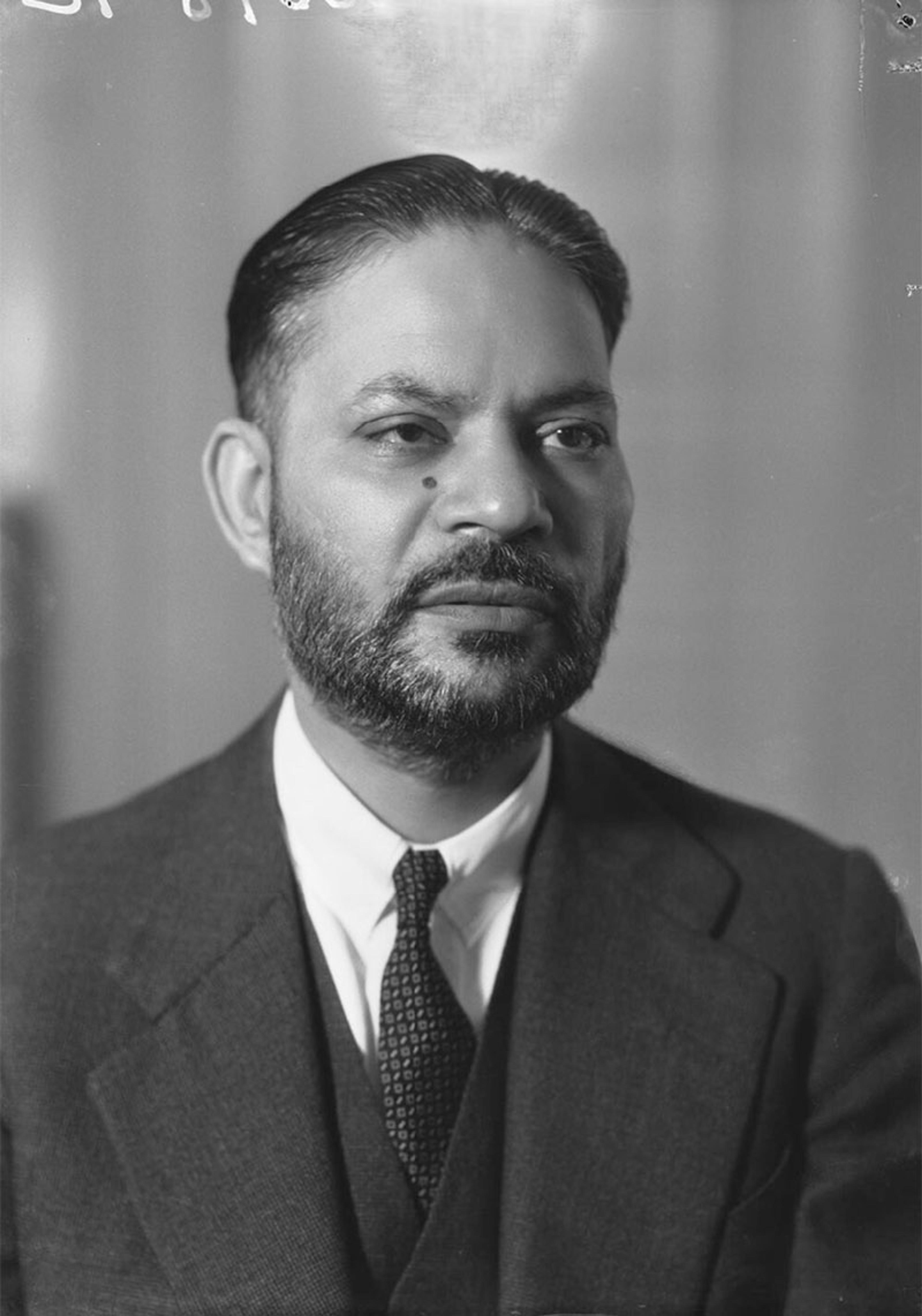 Black-and-white photo of a man in a suit with a beard and moustache, looking to the side with a neutral expression.