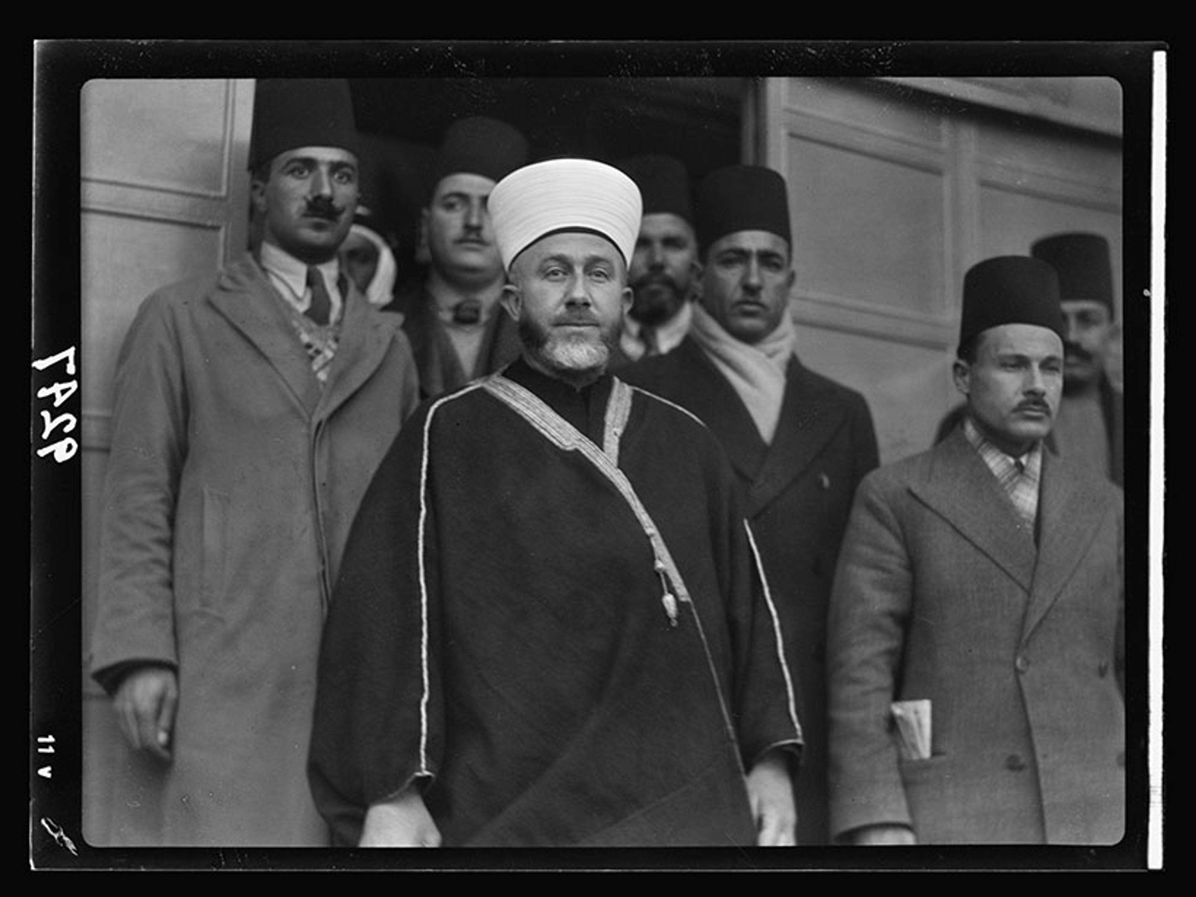 Black-and-white photo of a man in traditional attire with several men in suits and fezzes standing behind him.