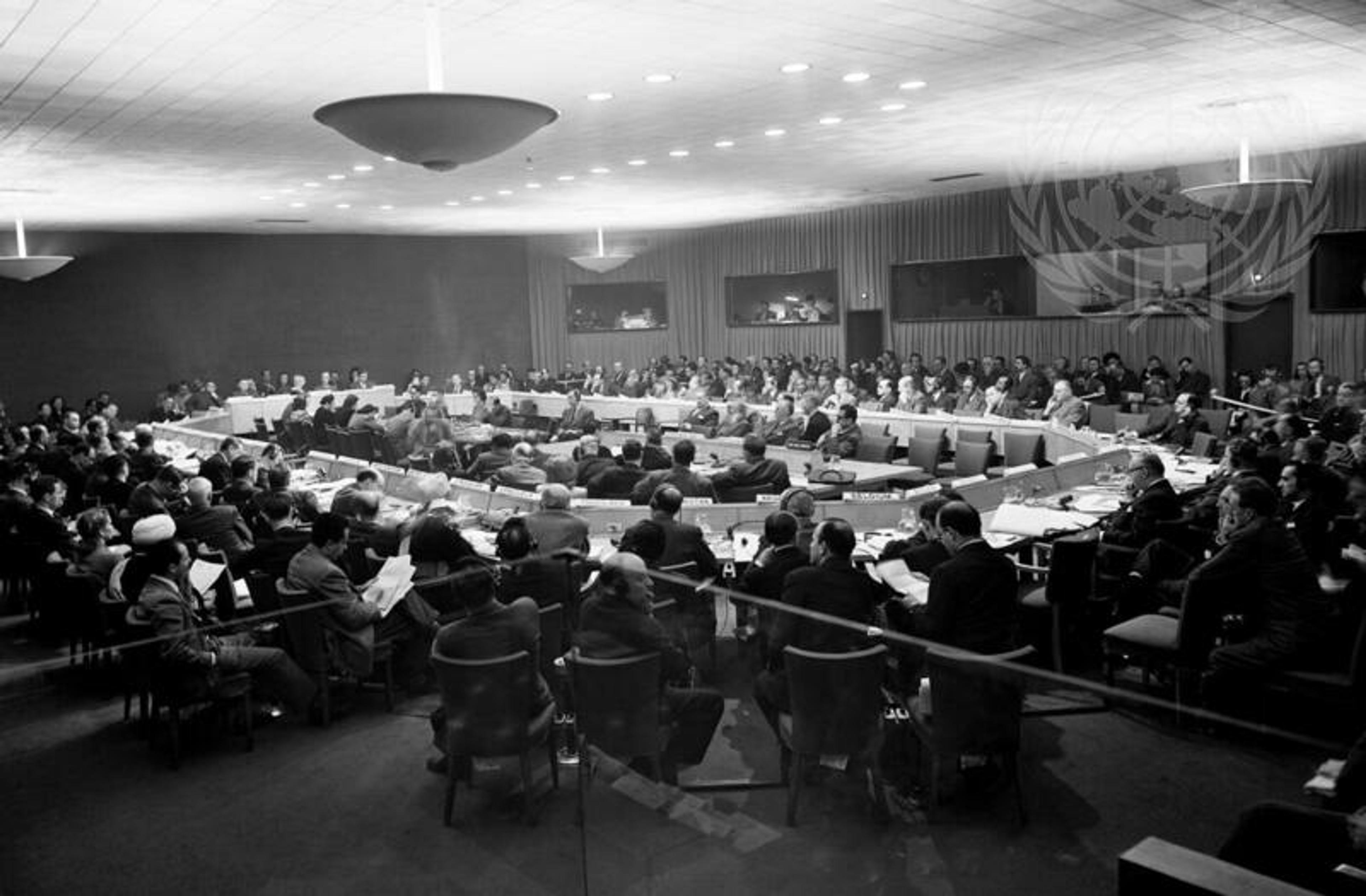 Black-and-white photo of a large meeting with delegates seated in a circular arrangement in a conference room.