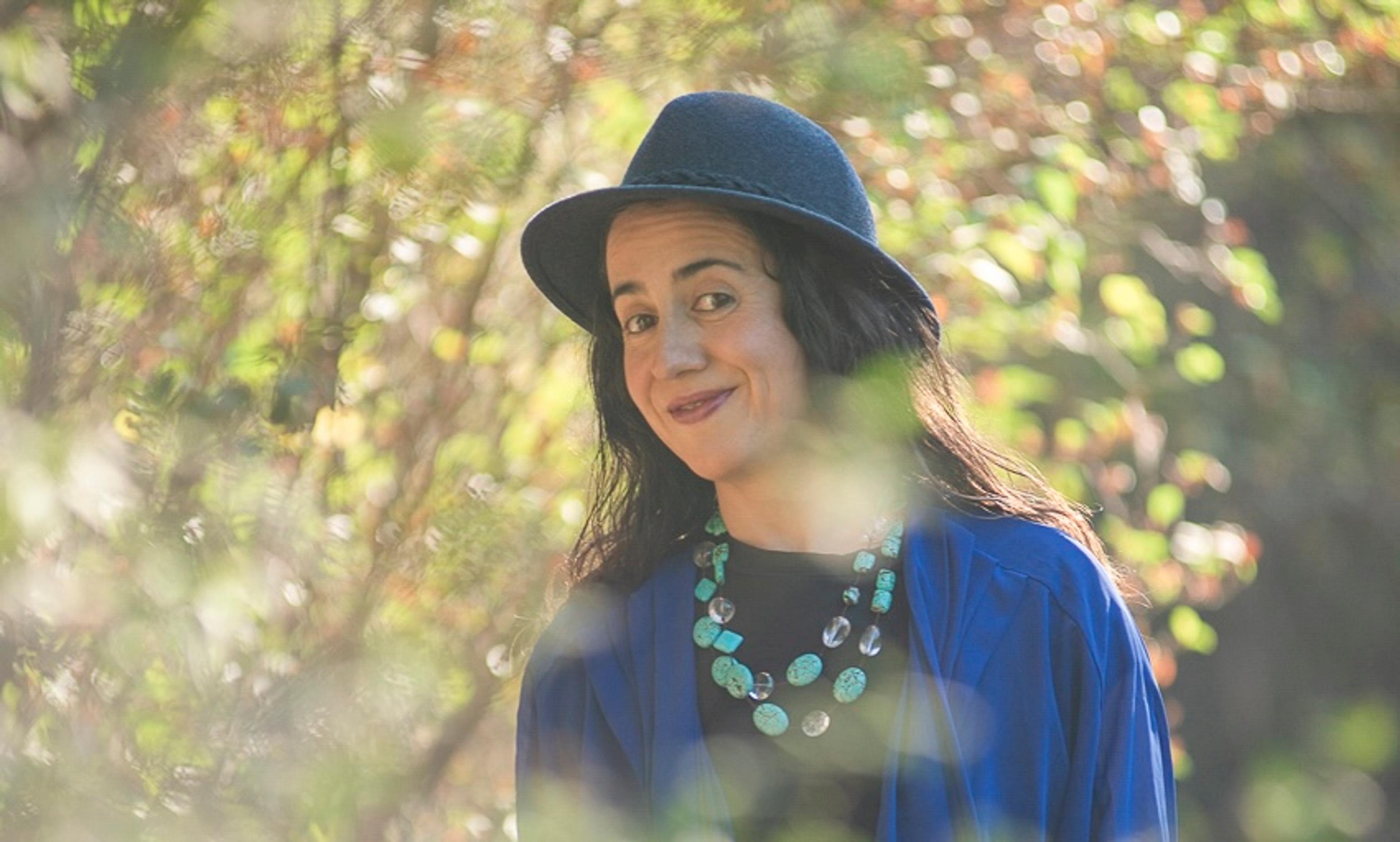 Photo of a smiling person in a hat and blue necklace standing in a sunlit, leafy garden setting.