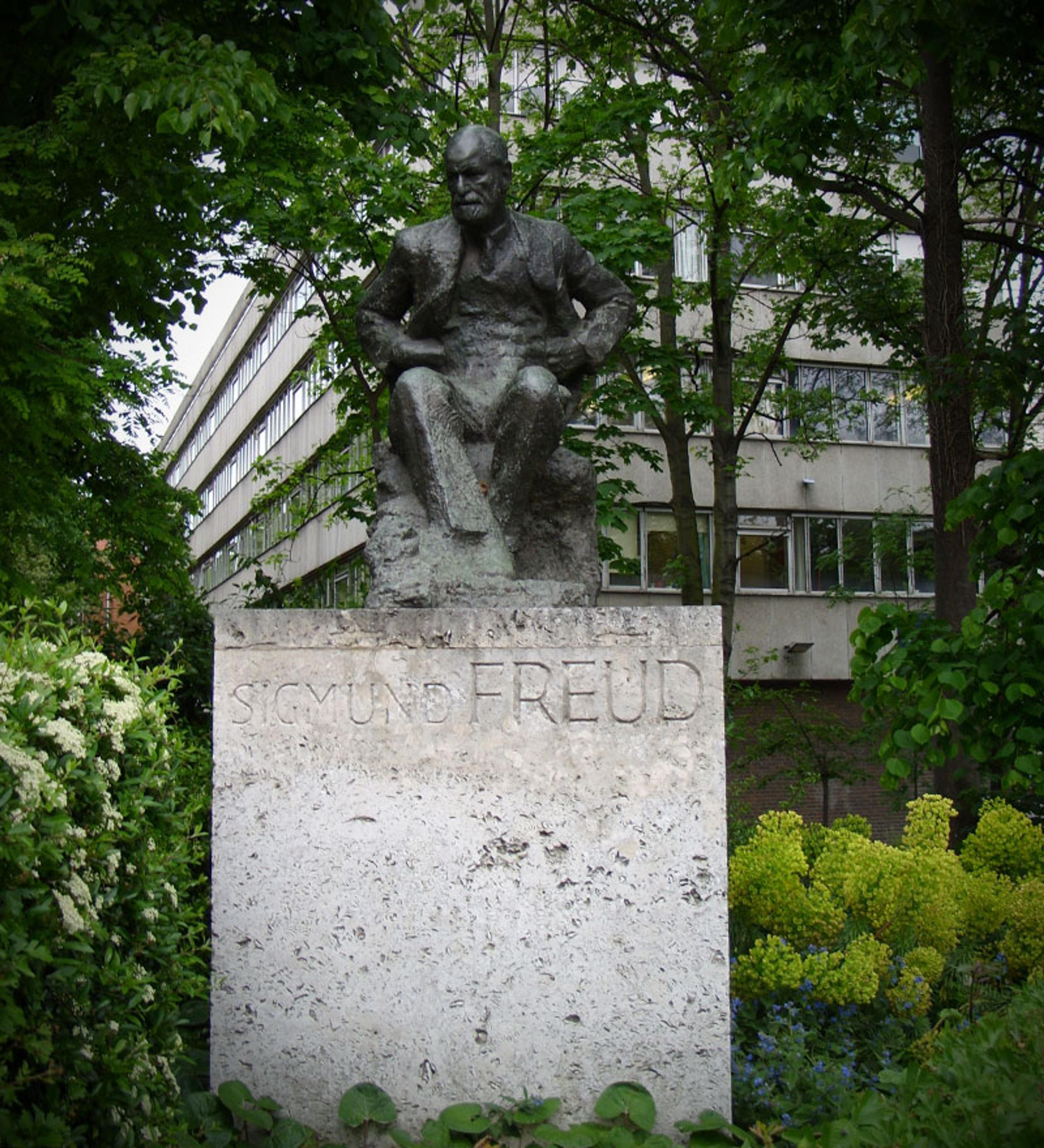 Photo of a Sigmund Freud statue on a stone pedestal surrounded by trees and greenery near a building.