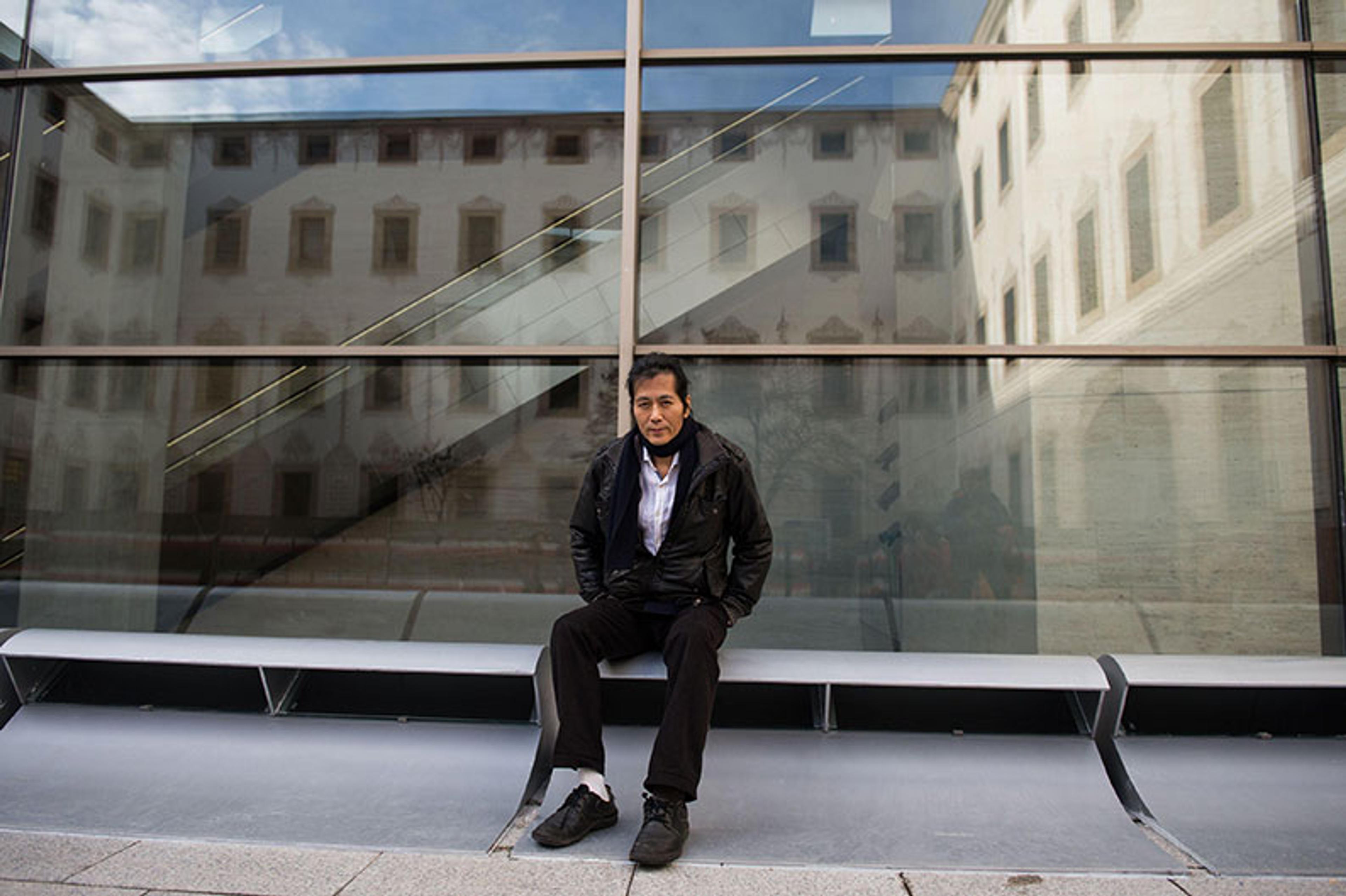 Photo of a man sitting on a bench in front of a glass building facade reflecting an adjacent structure.