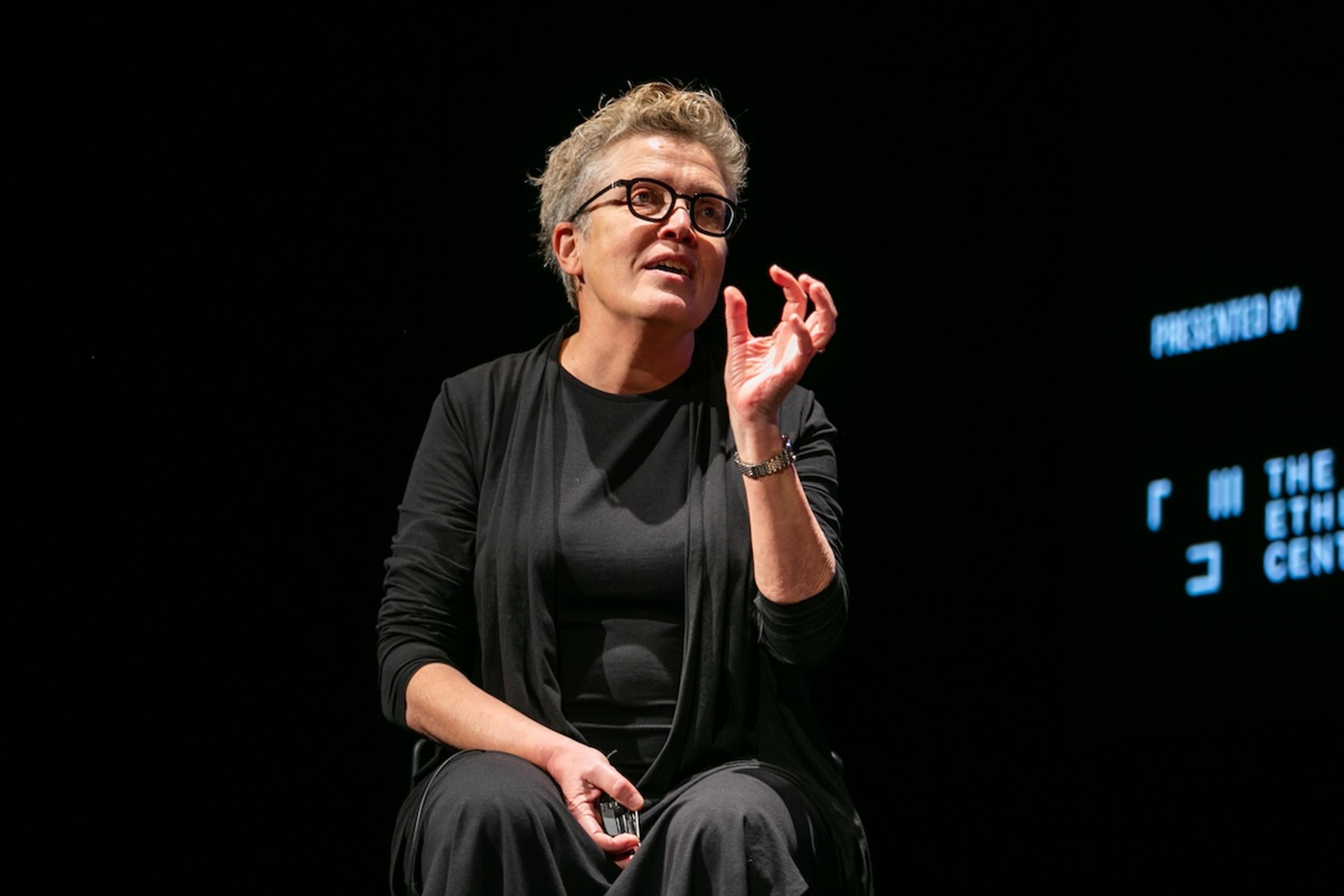 Photo of a person speaking on stage against a dark background wearing glasses and black attire gesturing with one hand.