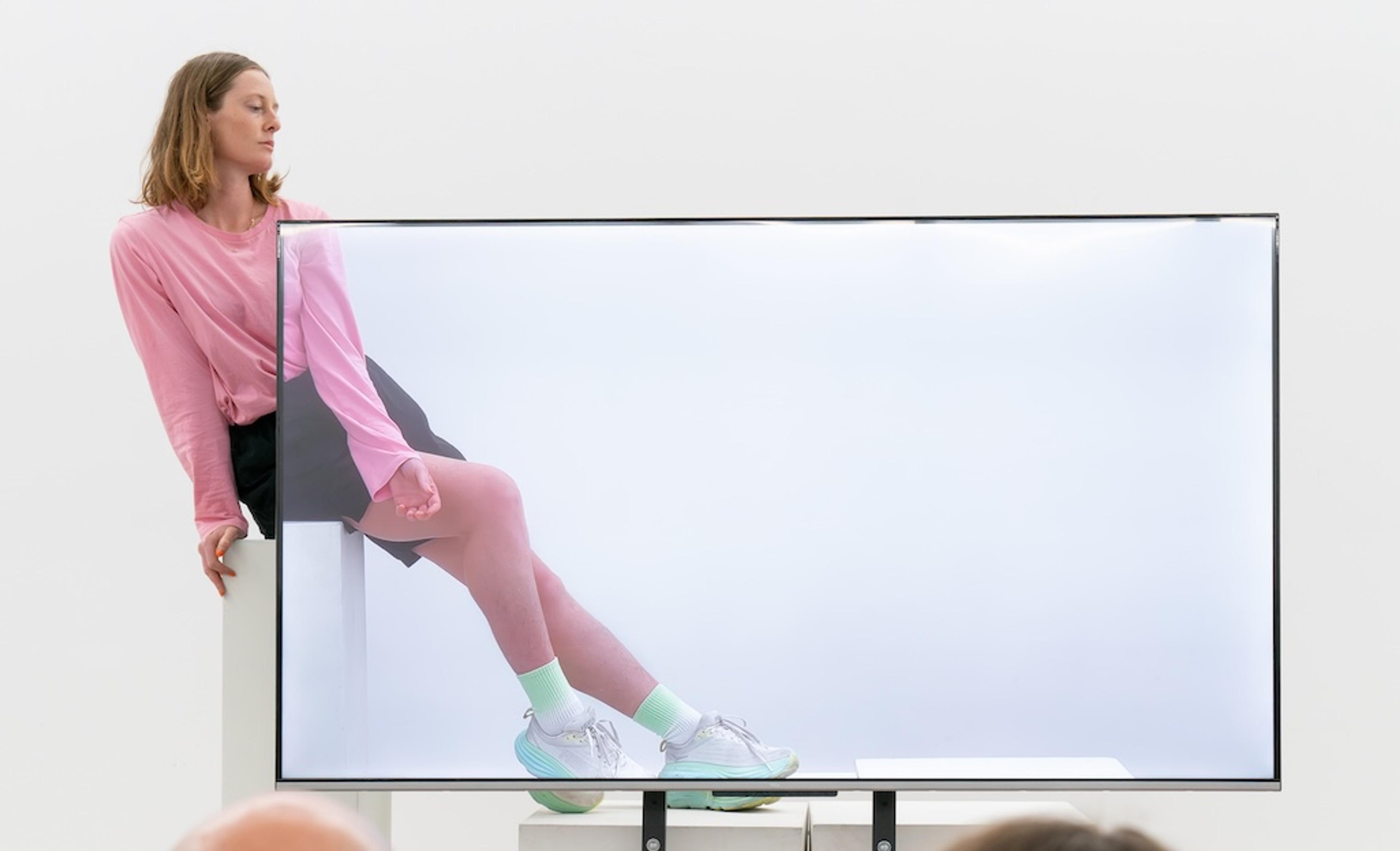 Photo of a woman in pink leaning on a white pedestal through a transparent screen, wearing a skirt and trainers.