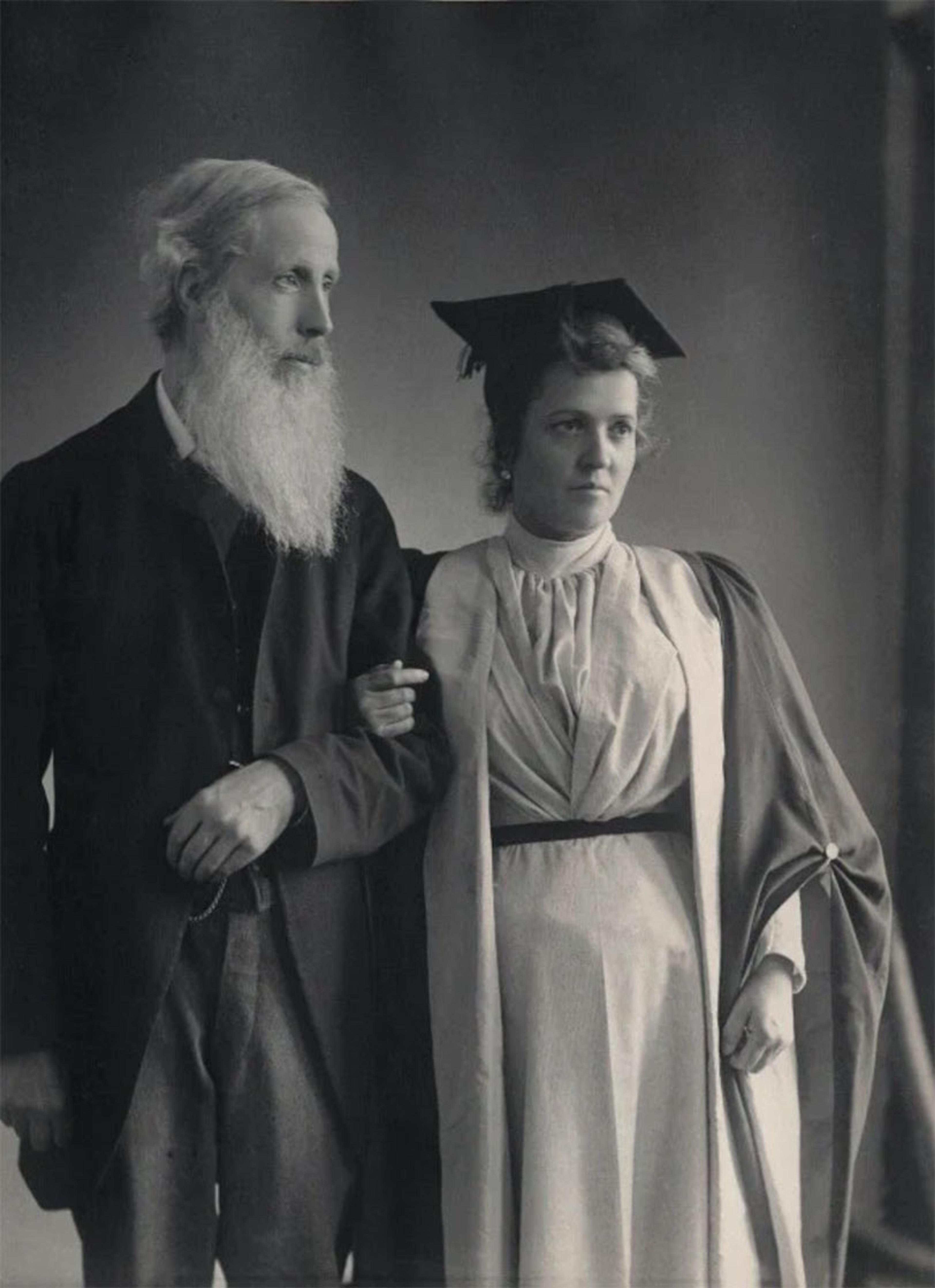 Black and white photo of a bearded man arm-in-arm with a woman in a mortarboard cap and gown, vintage style.