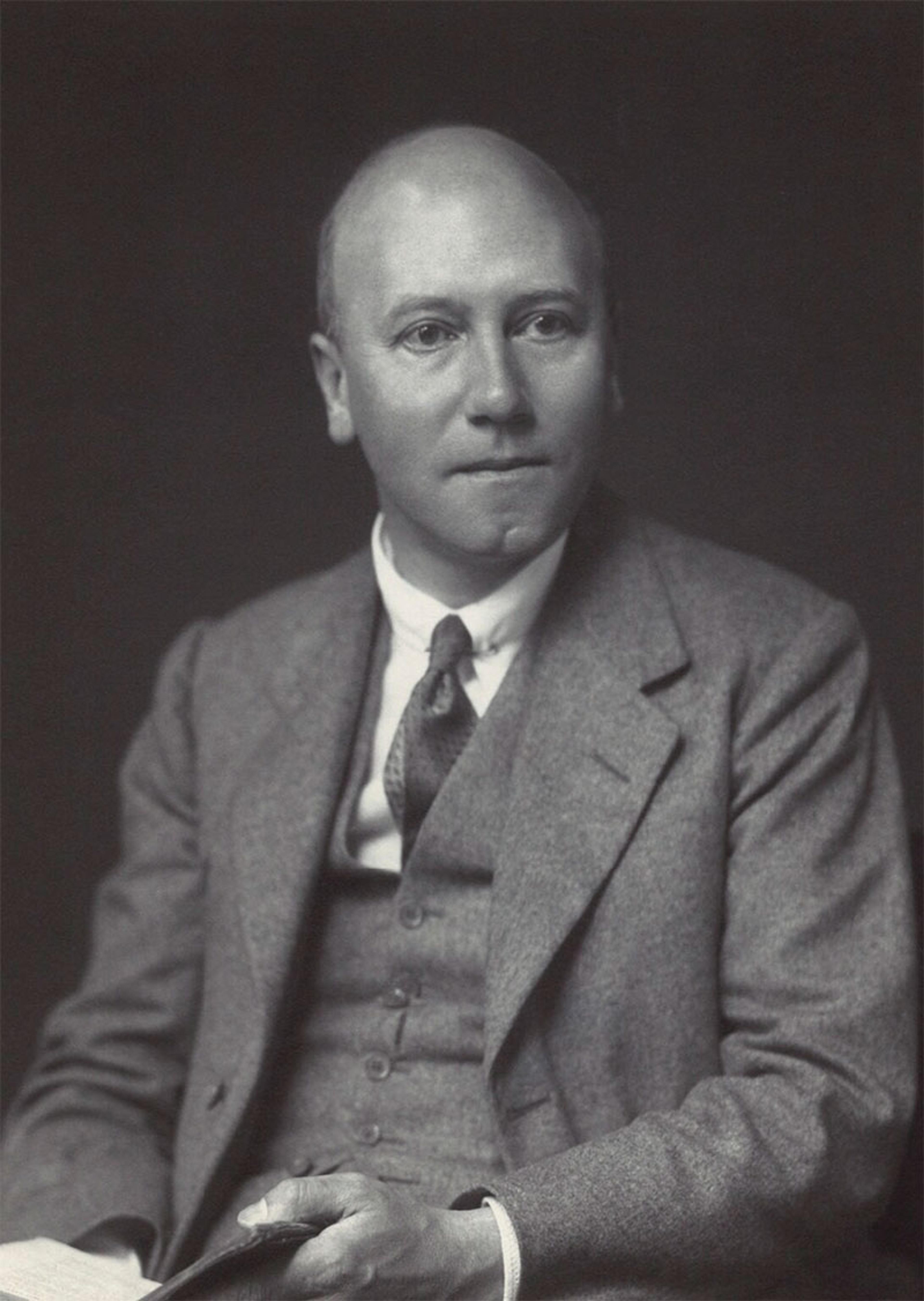 Black and white photo of a man in a suit holding a book sitting against a dark background.