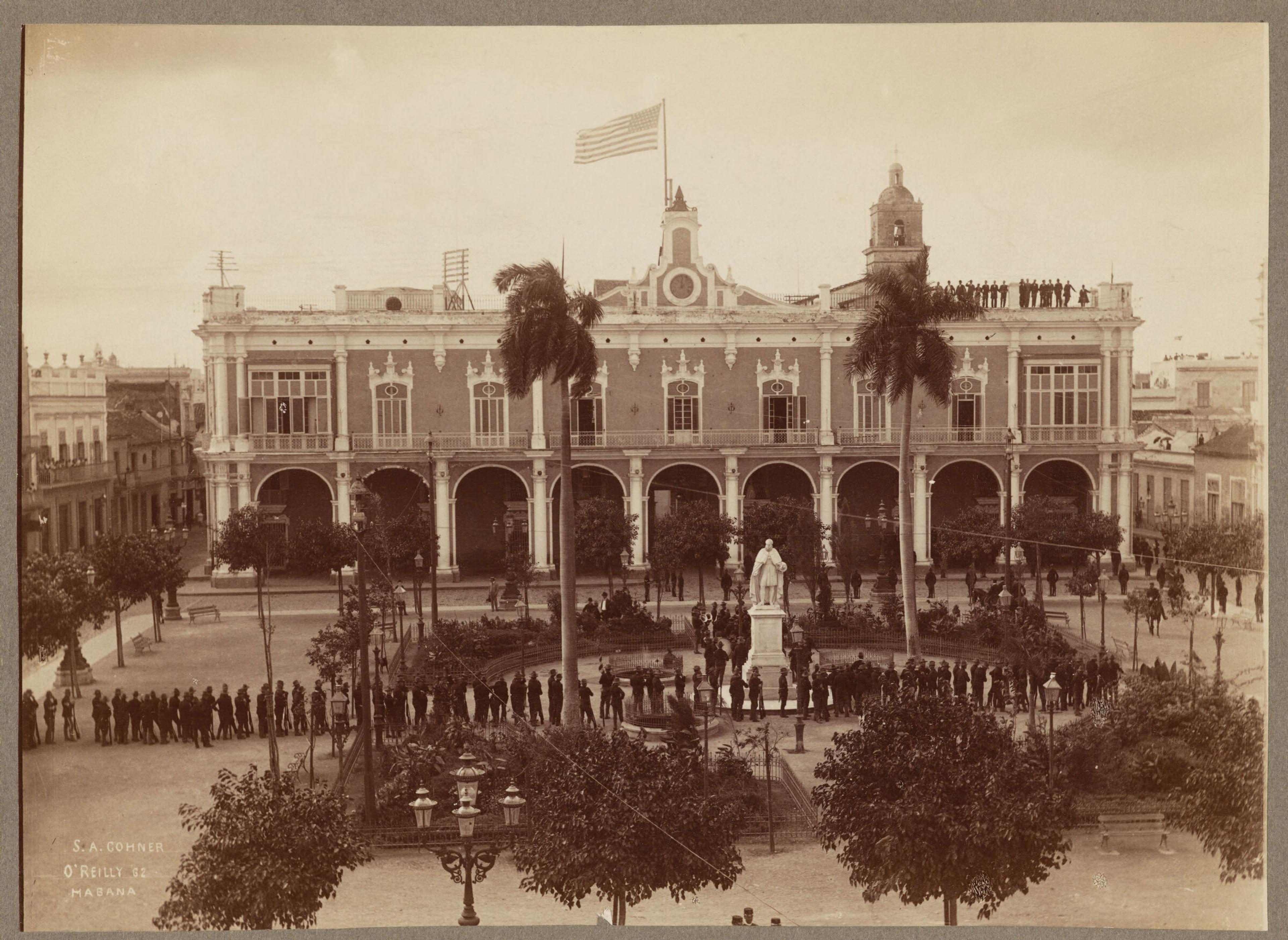 Sepia-toned photo of a colonial building with arches, palm trees and a statue in the foreground plaza.