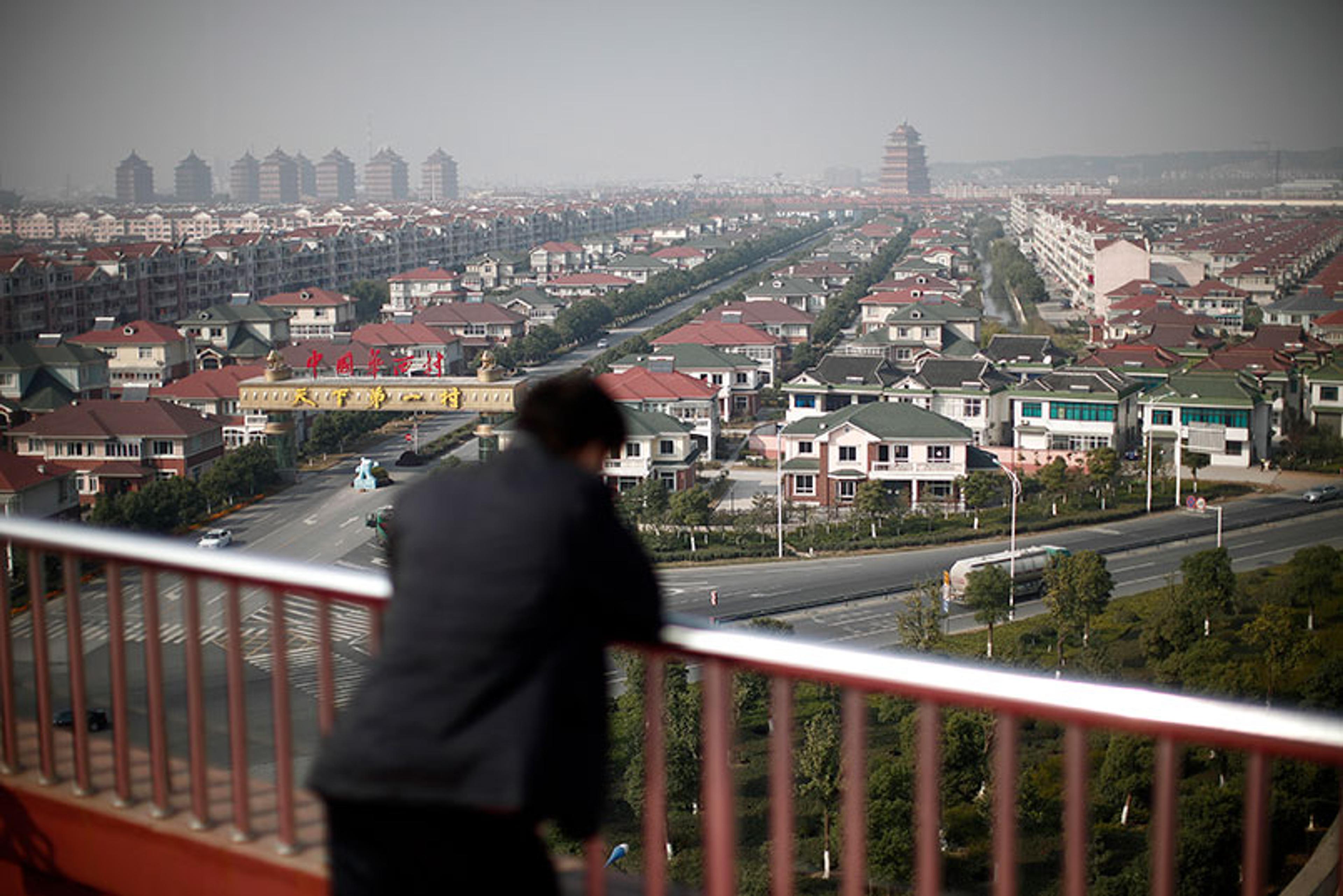 A person overlooking a vast cityscape with houses, a long road and pagoda in the distance under a hazy sky.