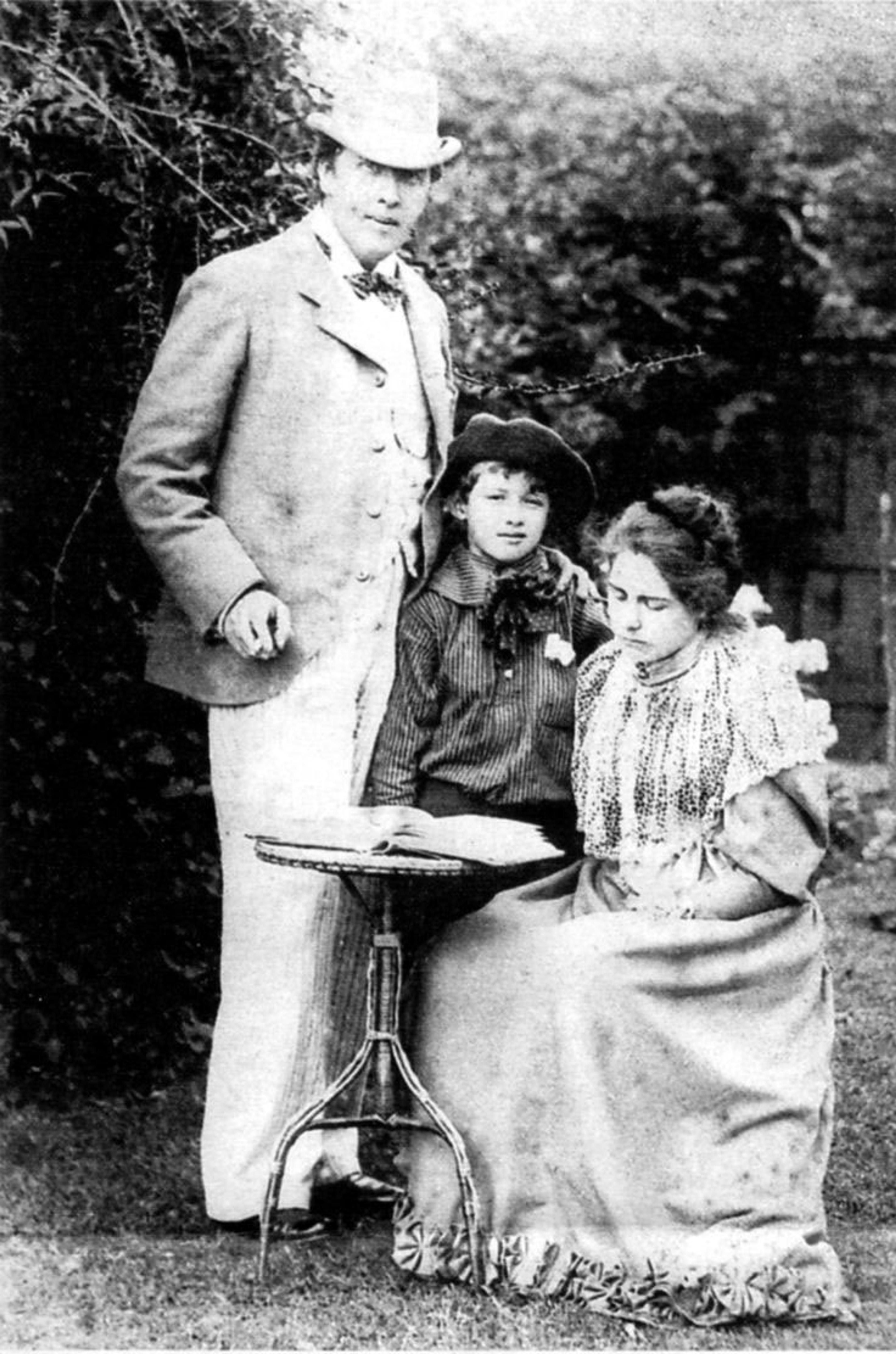 Vintage photo of a man in a suit, a seated woman reading, and a child with a hat in a garden setting.