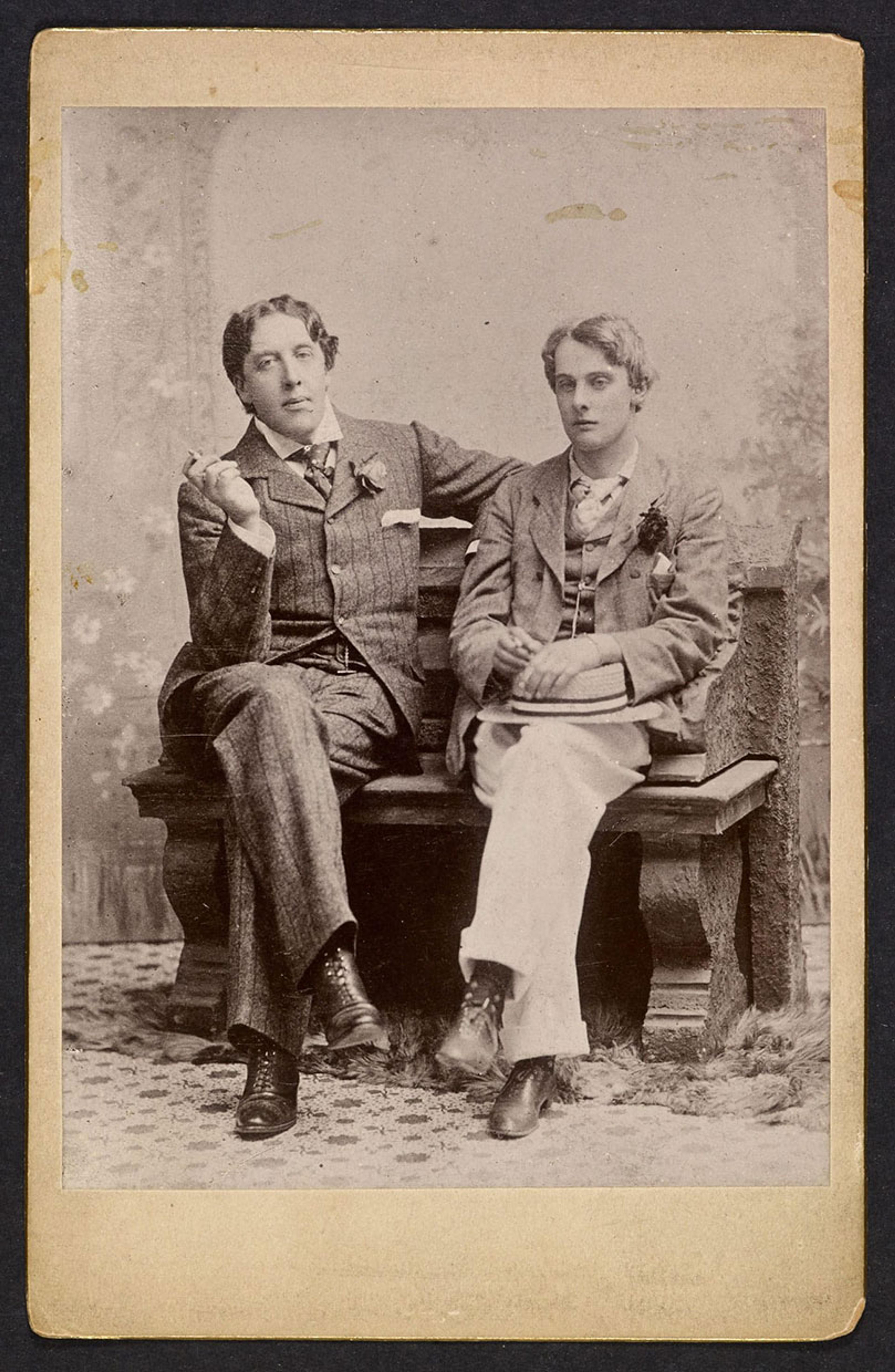 Vintage black and white photo of two men in suits on a bench with a neutral backdrop.