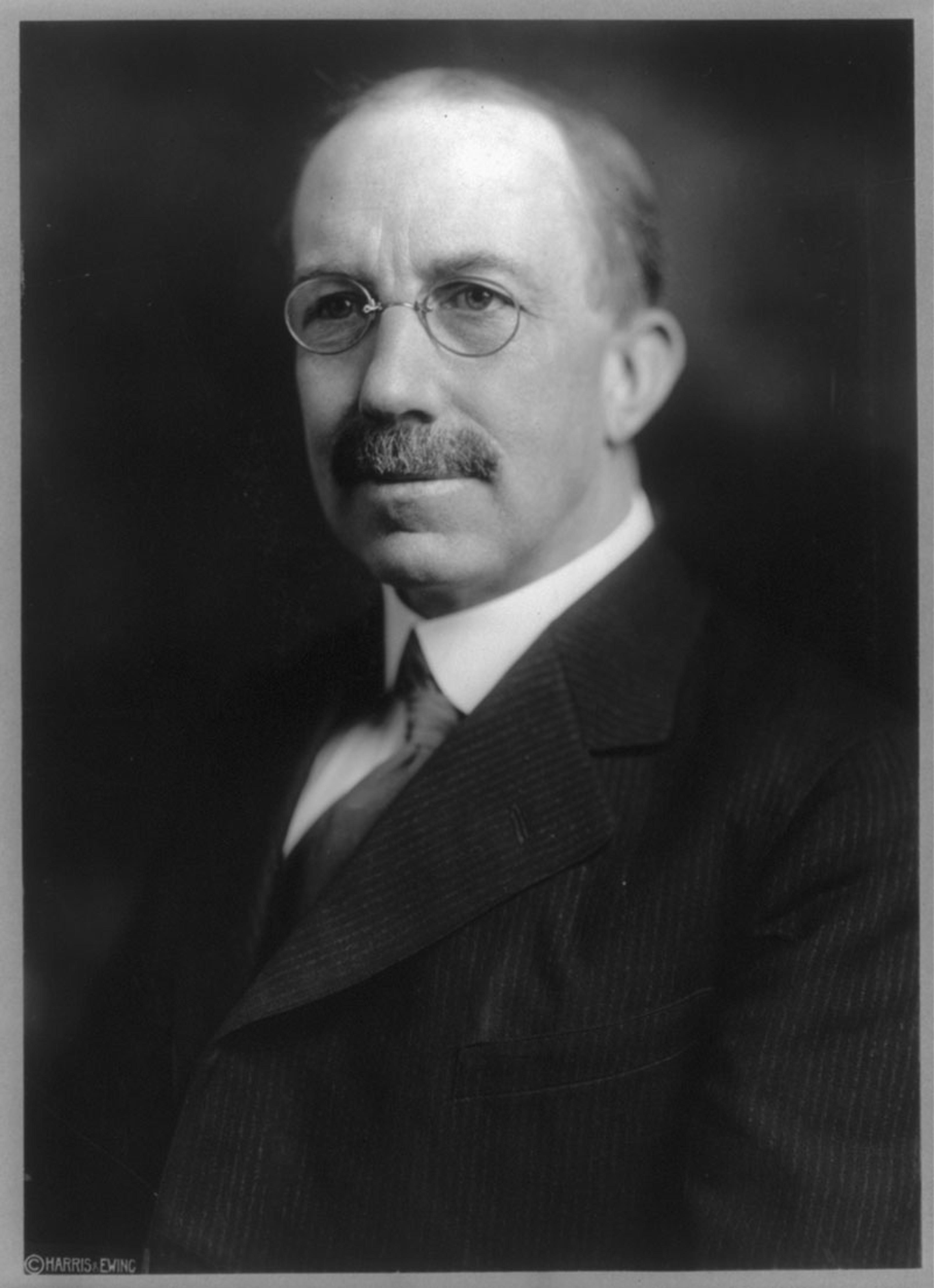 Black and white photo of an older man with glasses and a moustache wearing a suit against a dark background.