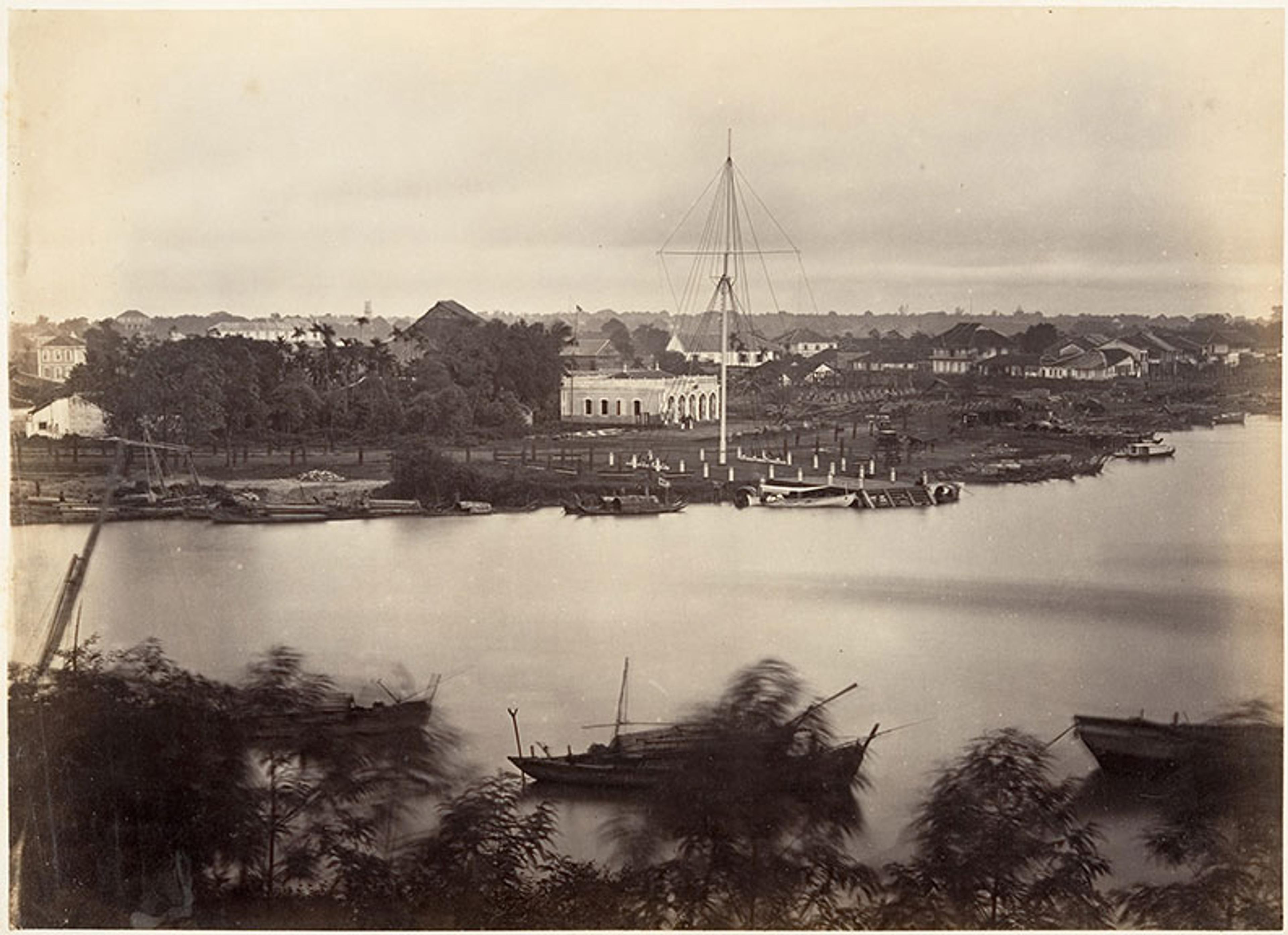 Historic photo of a waterfront scene with boats, a large building and trees in the background.