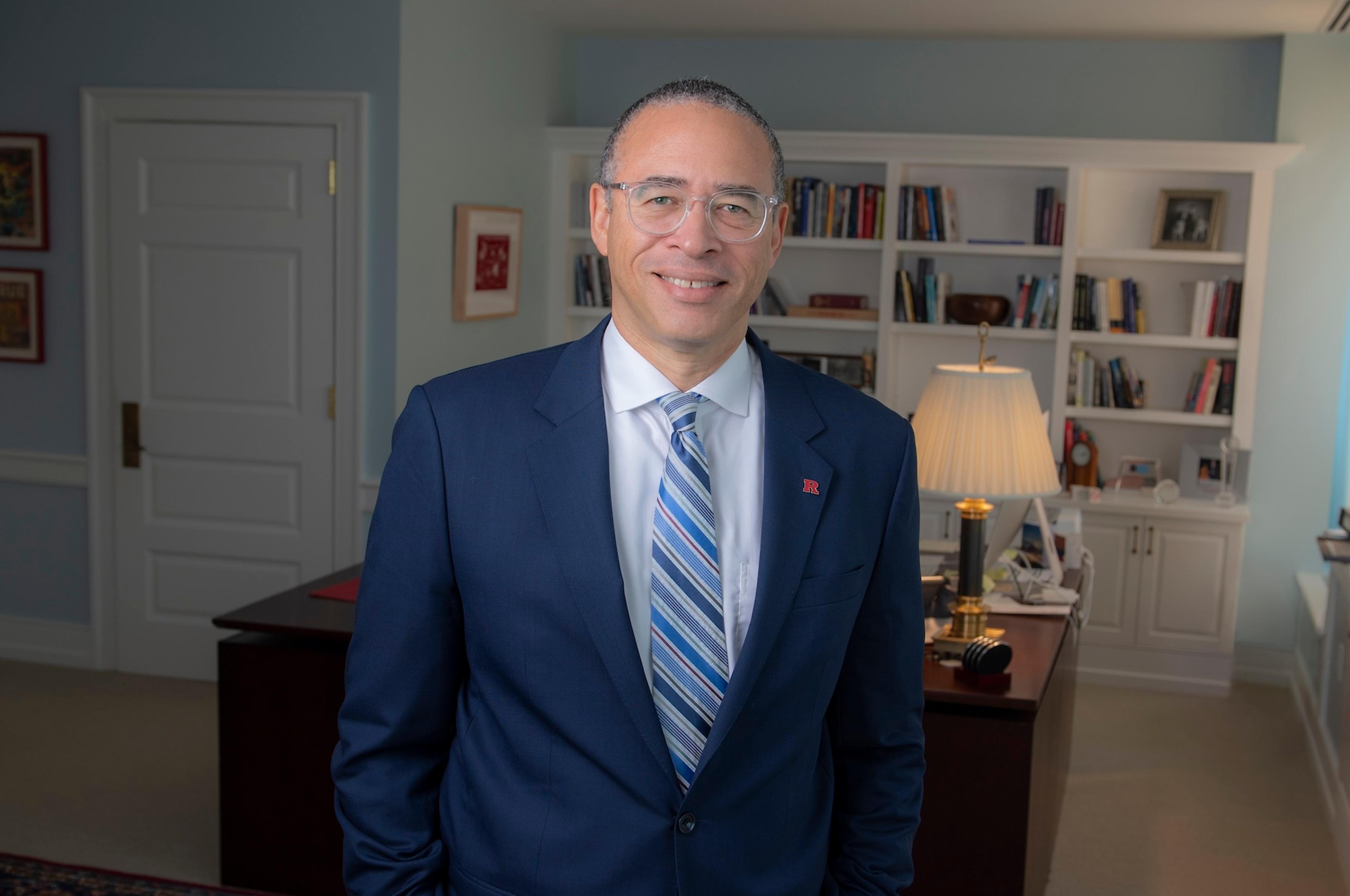 Photo of a person in a suit smiling in an office with a desk, lamp and bookshelves in the background.