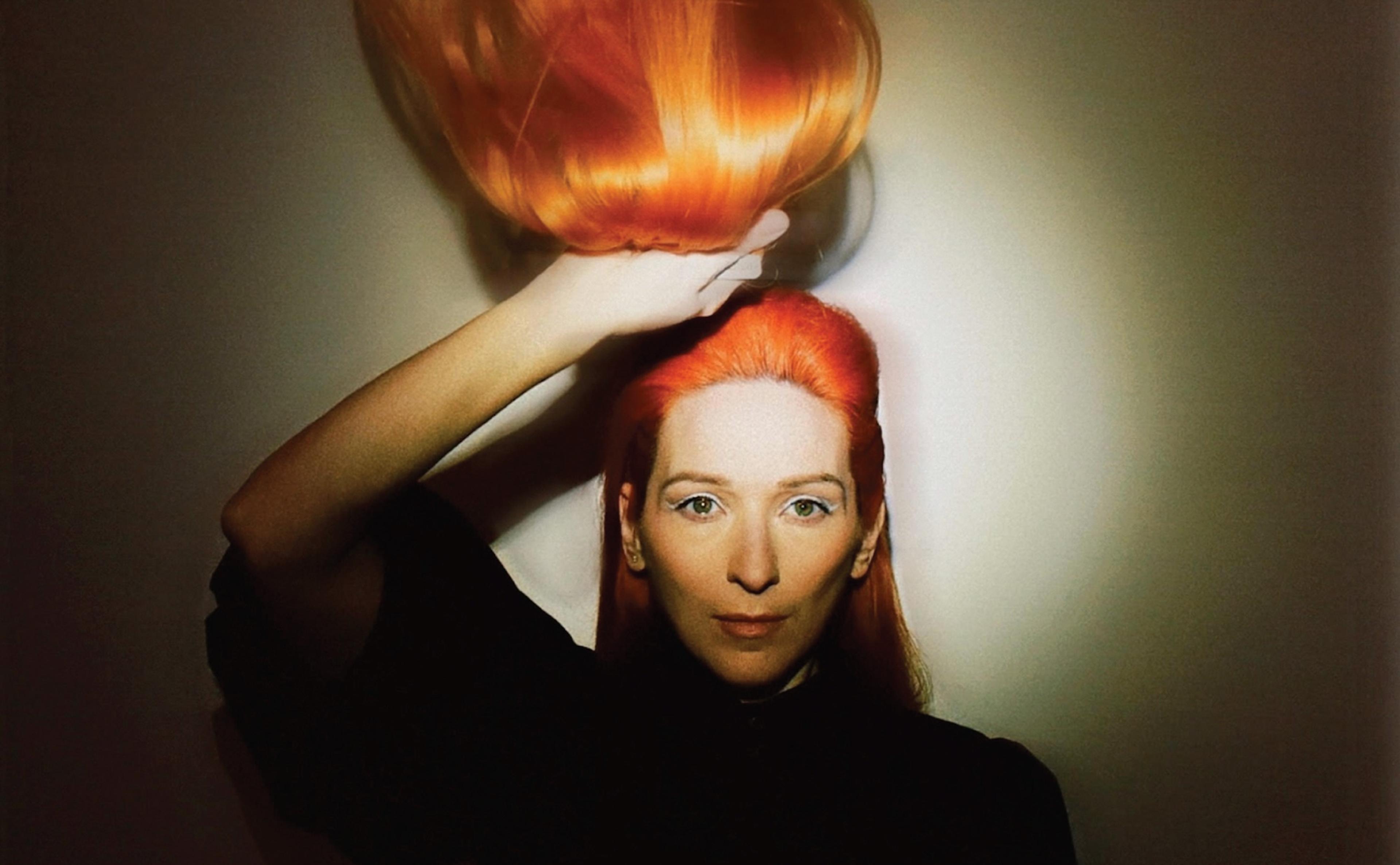 A photo of a person with bright red hair lifted in a hand, set against a soft shadowed background.