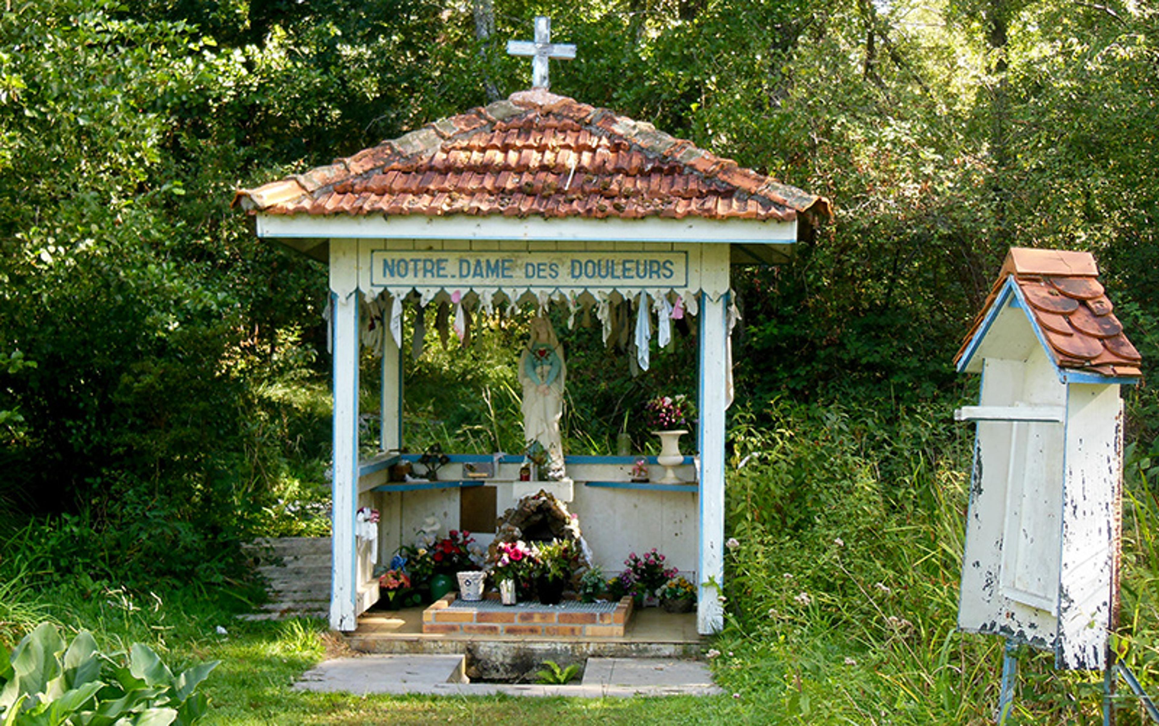 A small outdoor shrine titled “Notre Dame des Douleurs” with a statue of Mary, surrounded by greenery and flowers.