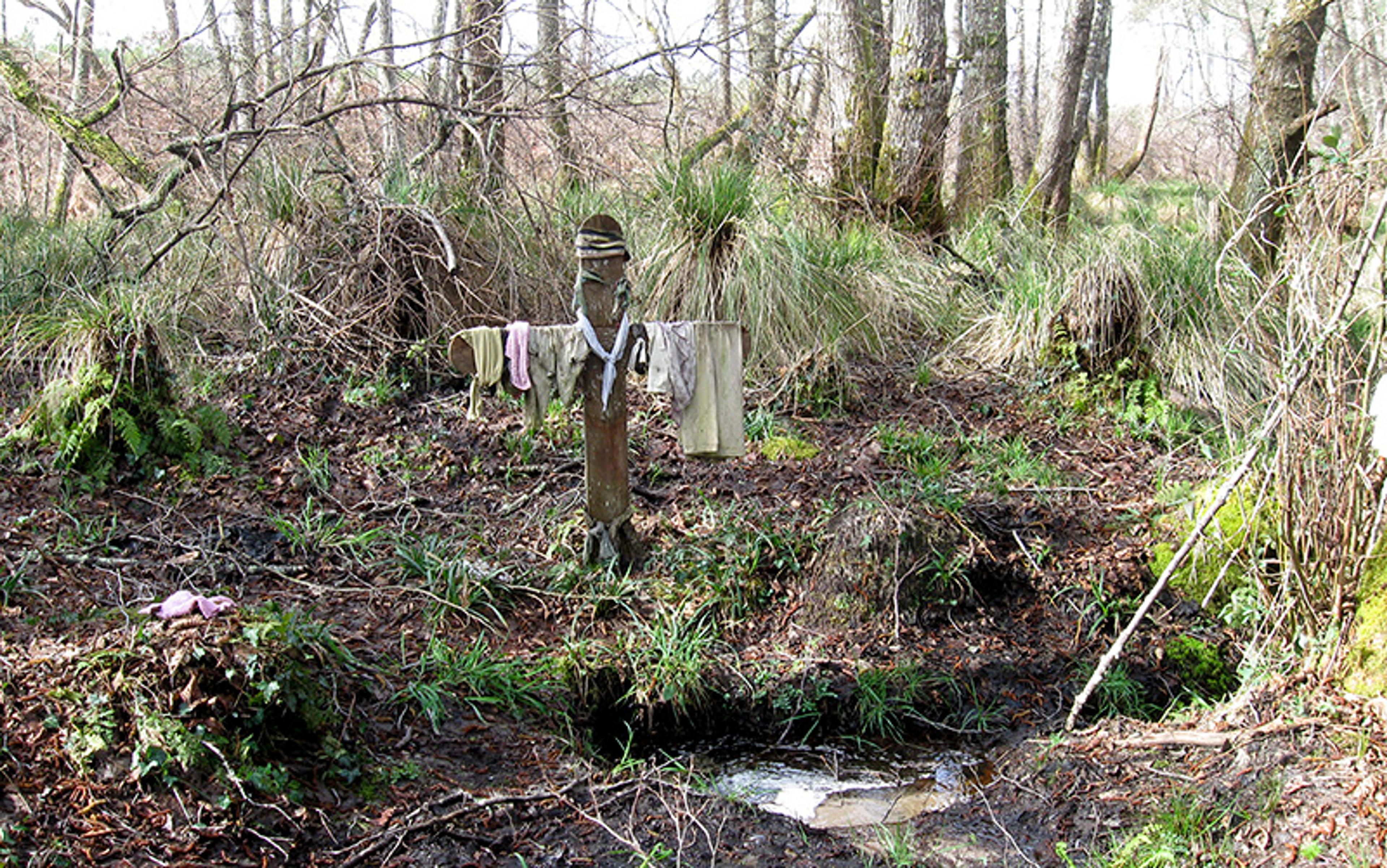 A woodland scene with a wooden cross dressed in hats and cloth, surrounded by trees and mossy undergrowth.