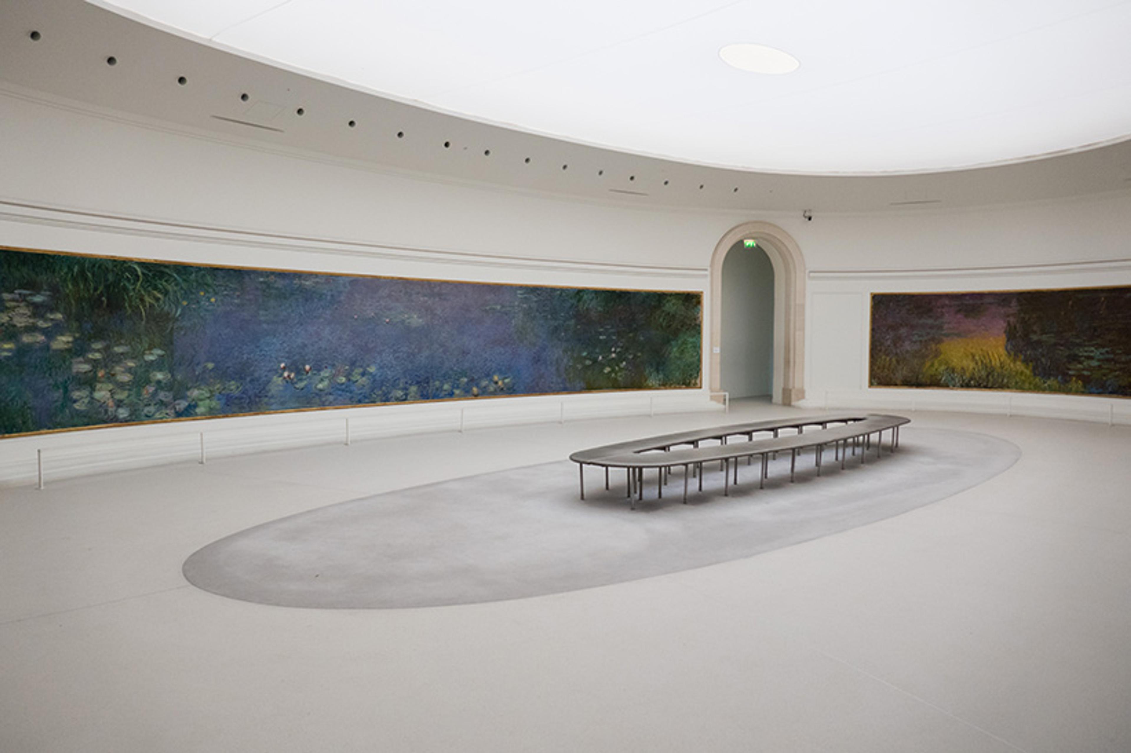 Photo of a white art gallery room with large Monet paintings on curved walls and a central oval bench under a skylight.