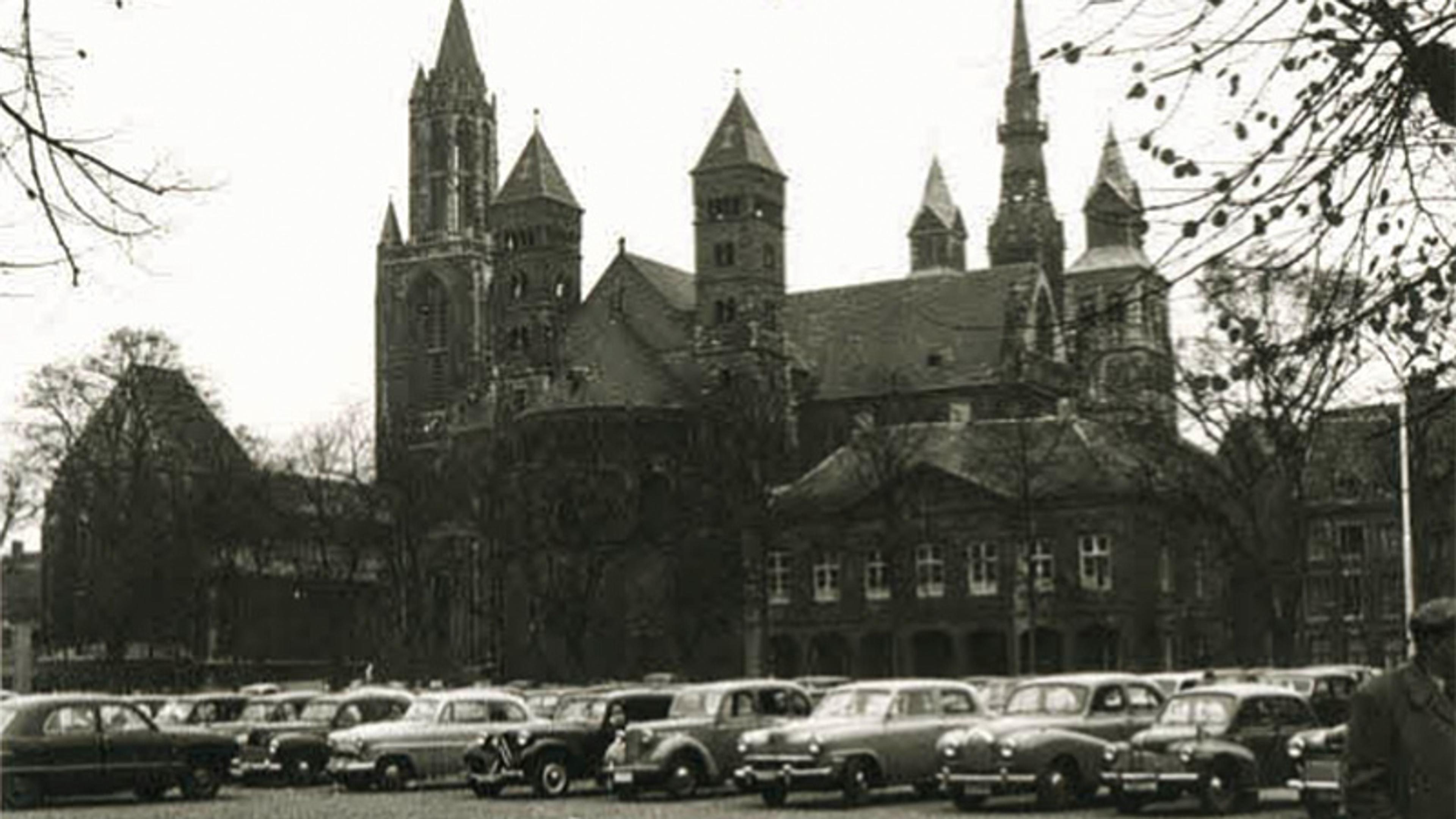 Black and white photo of a historic cathedral with multiple spires, cars parked in front, trees without leaves line the scene.