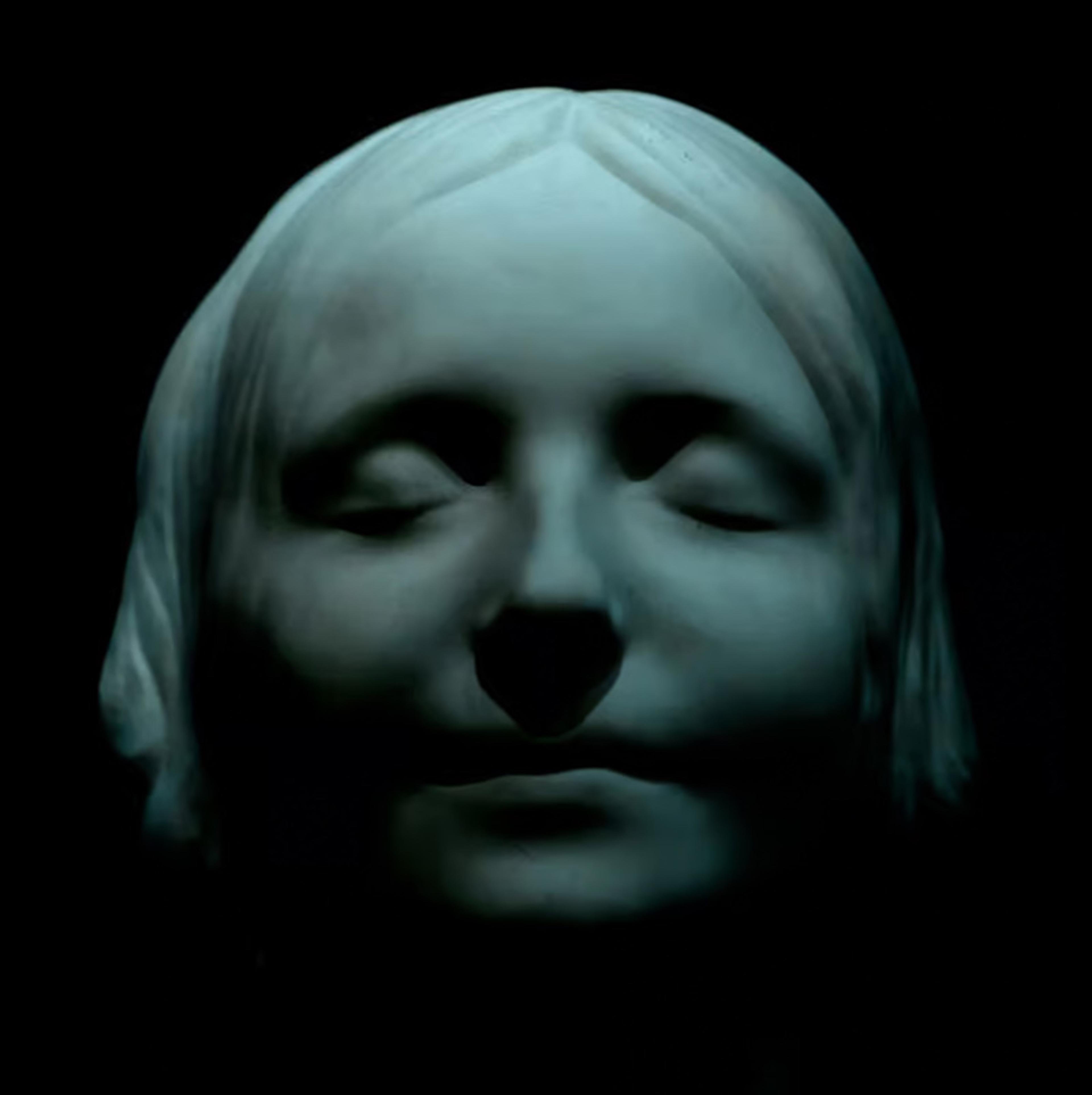 Sculpture of a serene face with closed eyes against a dark background lit in blue-green hues.