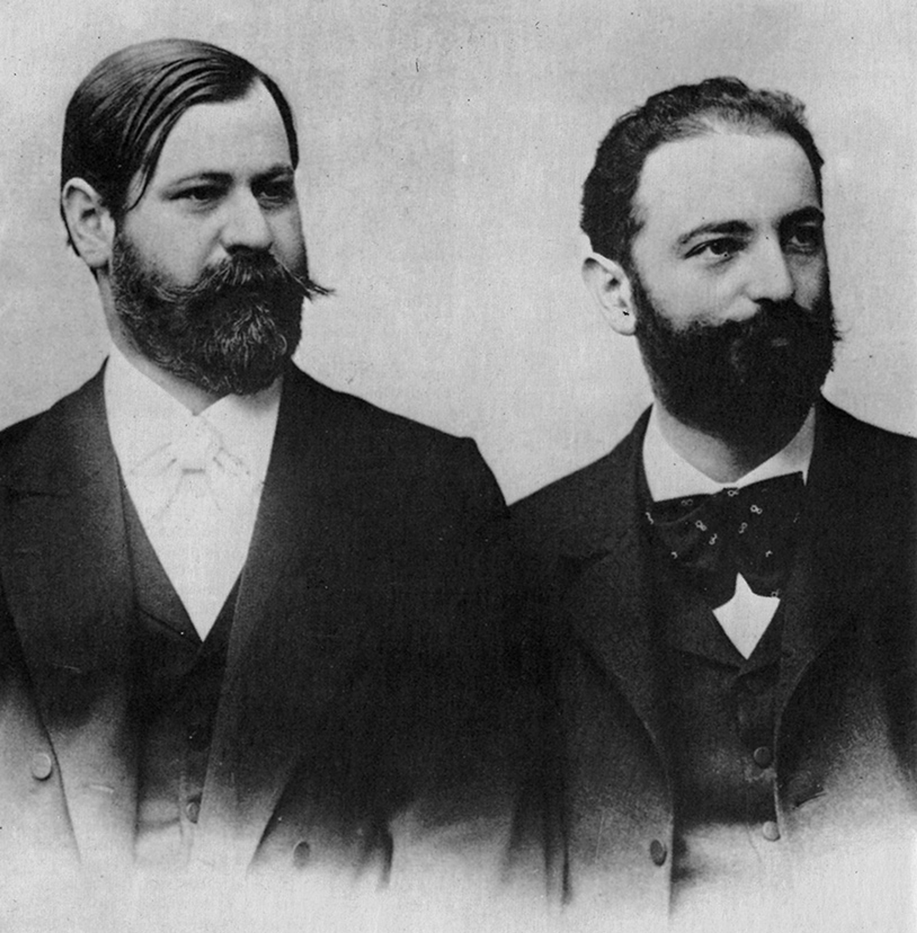 Vintage photo of two men in suits with beards, one with slicked hair and the other with curly hair, on a neutral background.