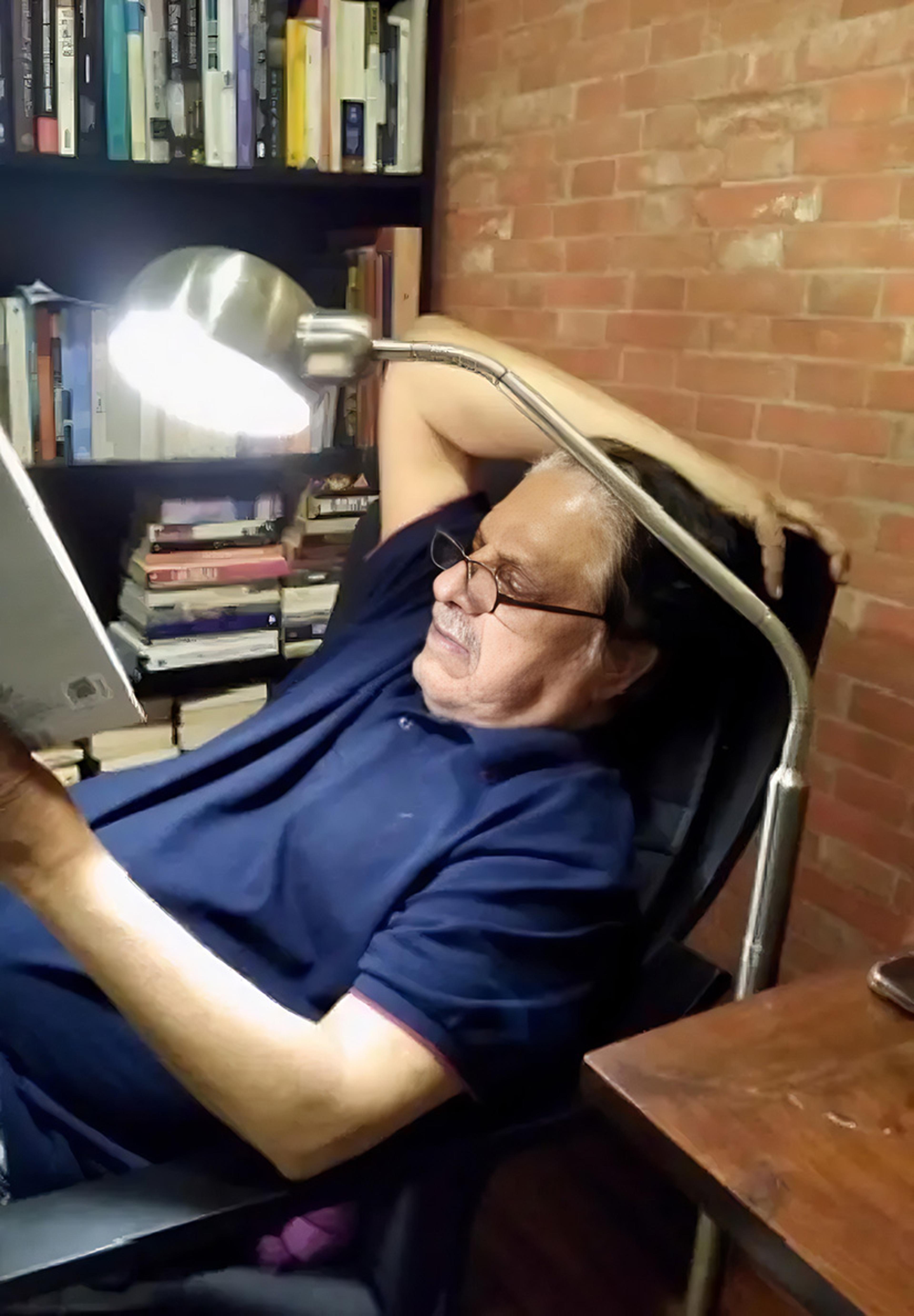 A man wearing glasses reading a book while leaning back in a chair with a lamp and bookshelves beside him.