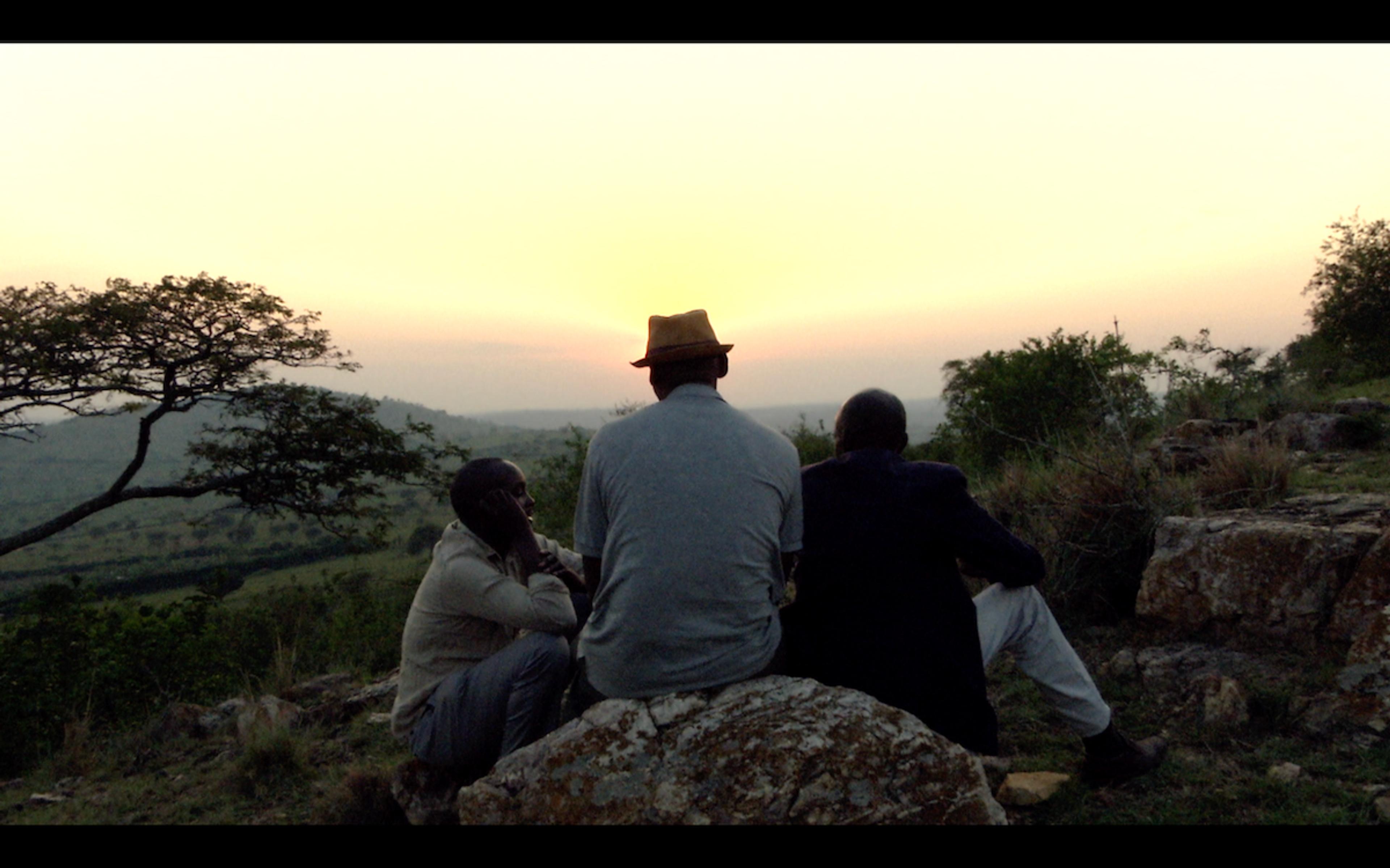 Photo of three people sitting on rocks watching a sunset over a scenic landscape with trees and hills in the distance.