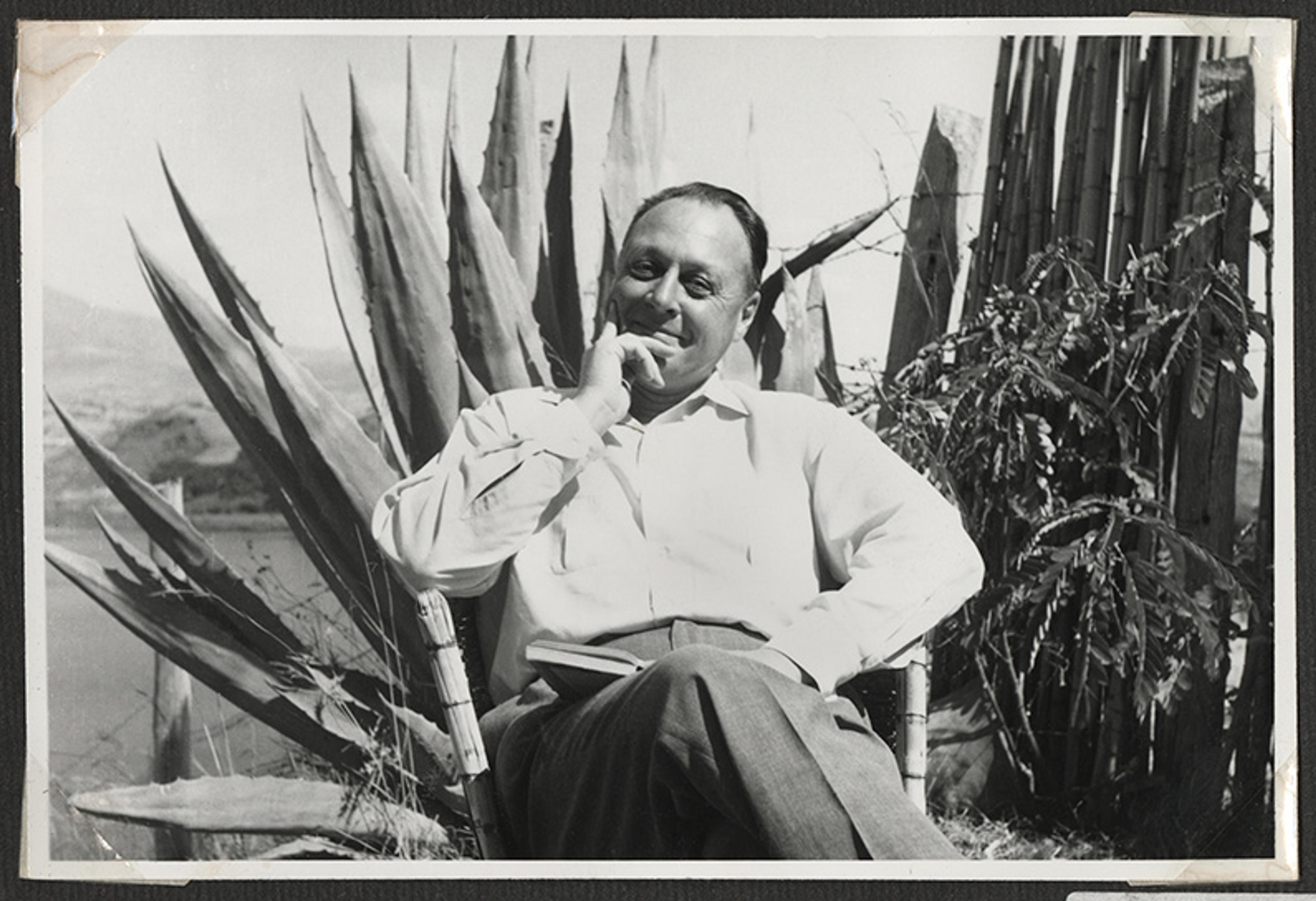 Black and white photo of a man in a white shirt sitting in a chair outdoors next to large agave plants, holding a book.