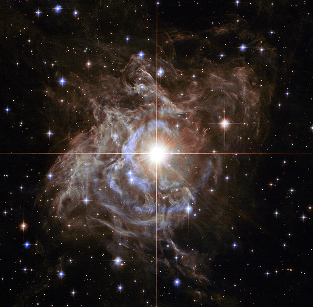 A luminous star at the centre of a swirling stellar nebula with bright stars scattered in the dark space background.