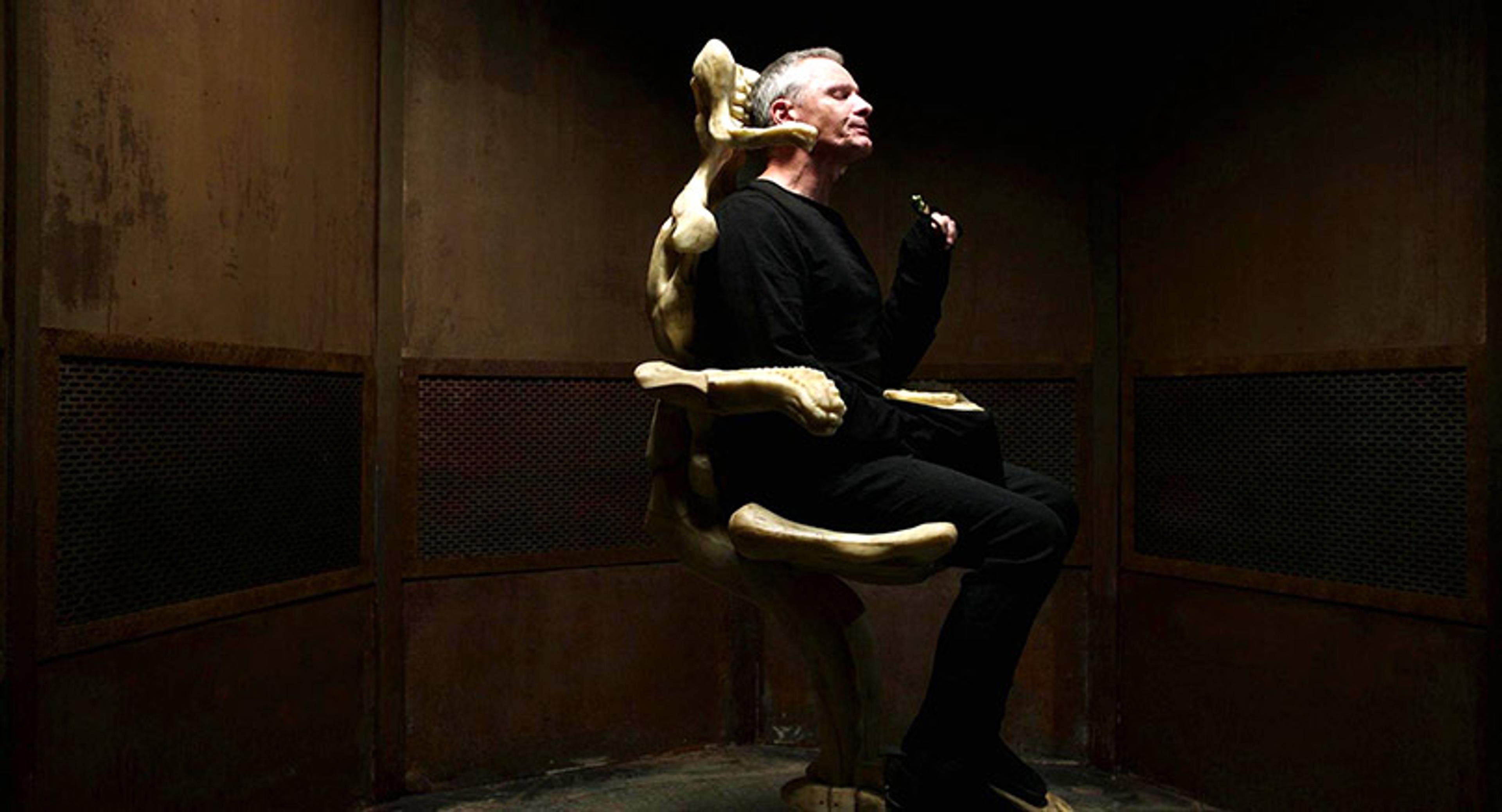 A man in dark clothes seated in a sculptural chair with skeletal forms against a dimly lit, industrial background.