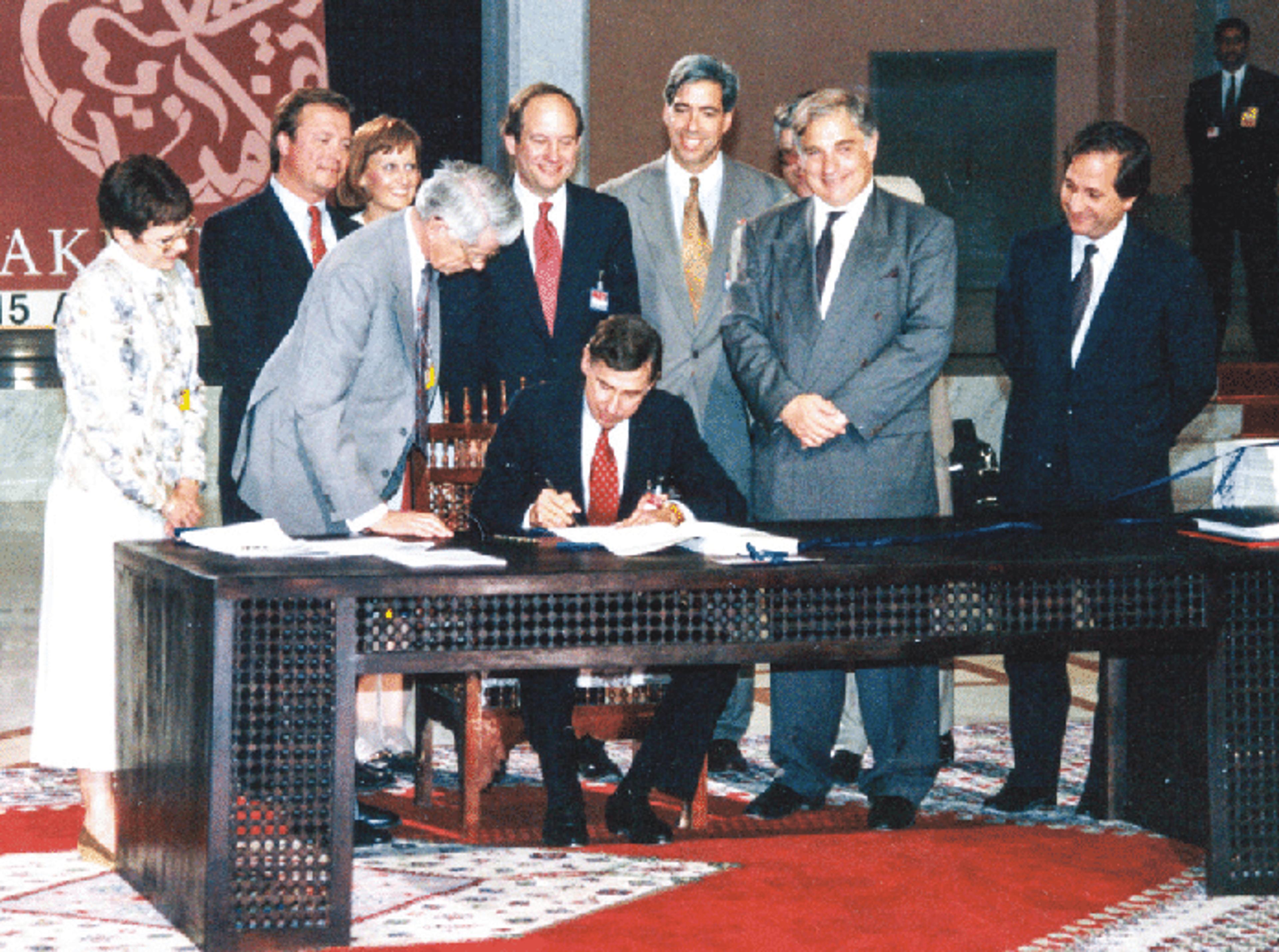 Photo of a man signing a document at a table with nine people standing behind him in a formal setting.