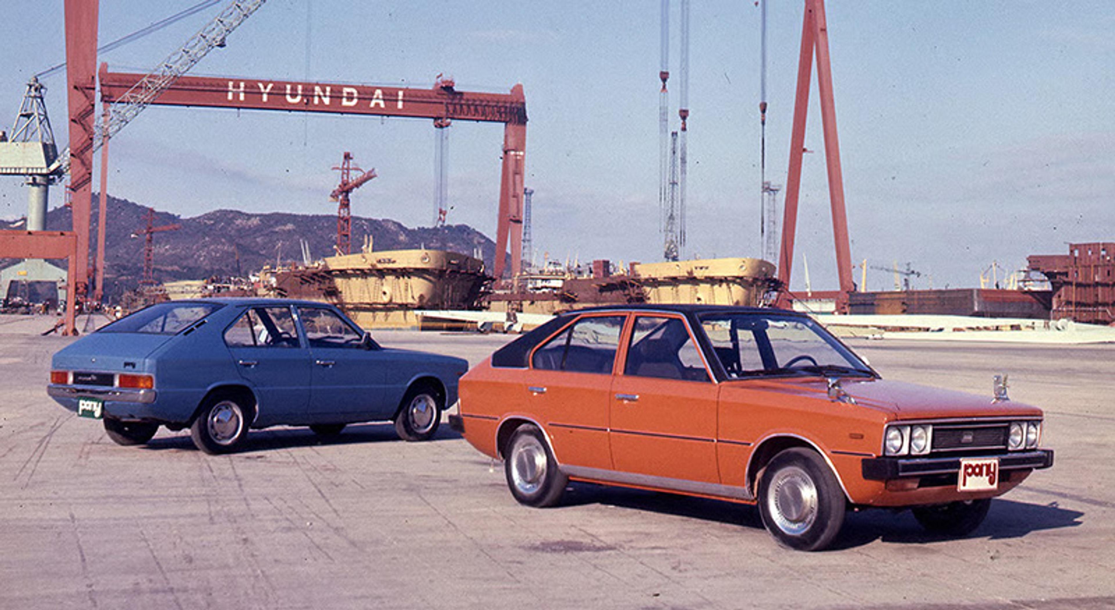 Two vintage cars, orange and blue, parked at an industrial port with a Hyundai crane and mountains in the background.