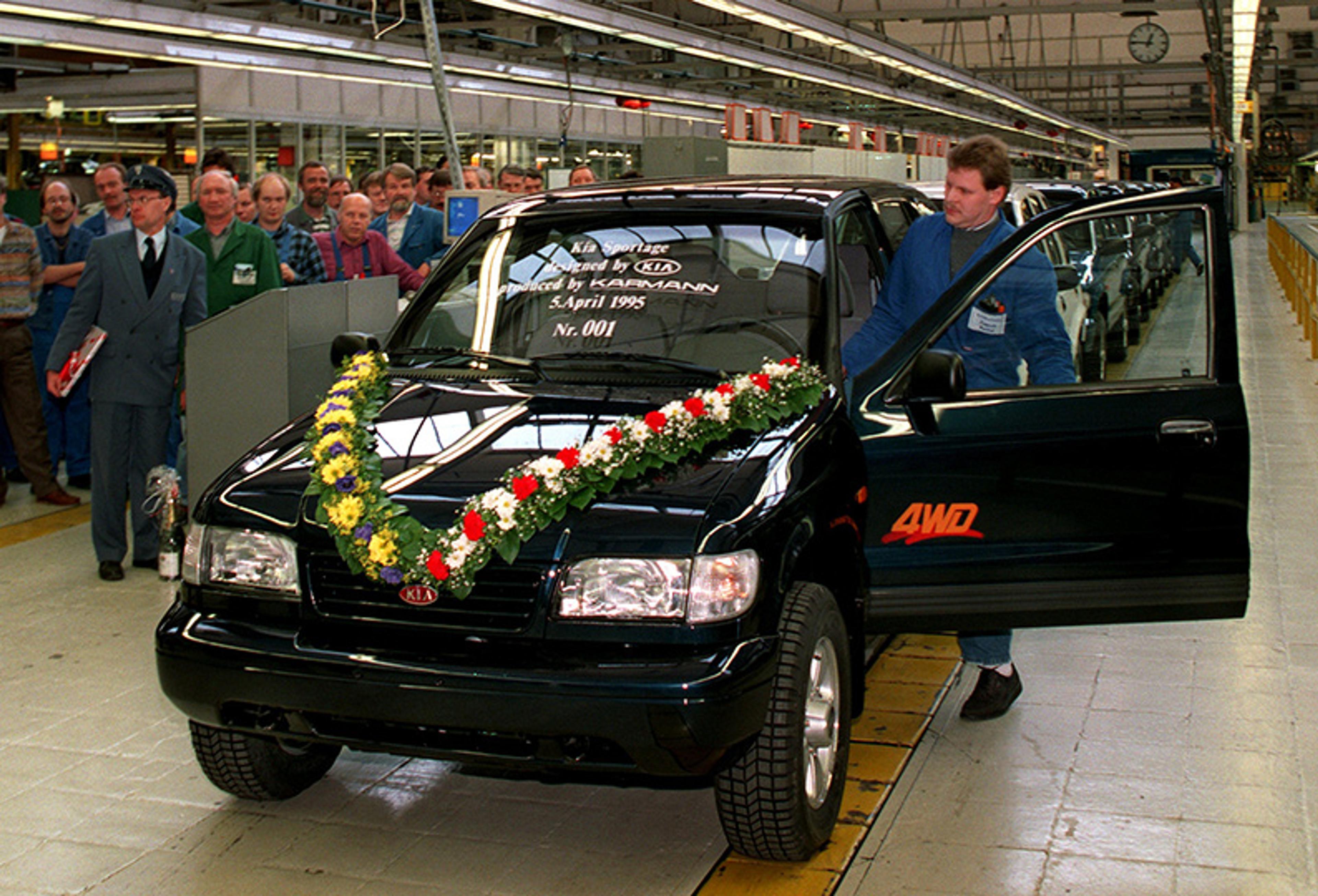 A dark SUV with a floral garland in a factory, surrounded by people, marking its production completion.