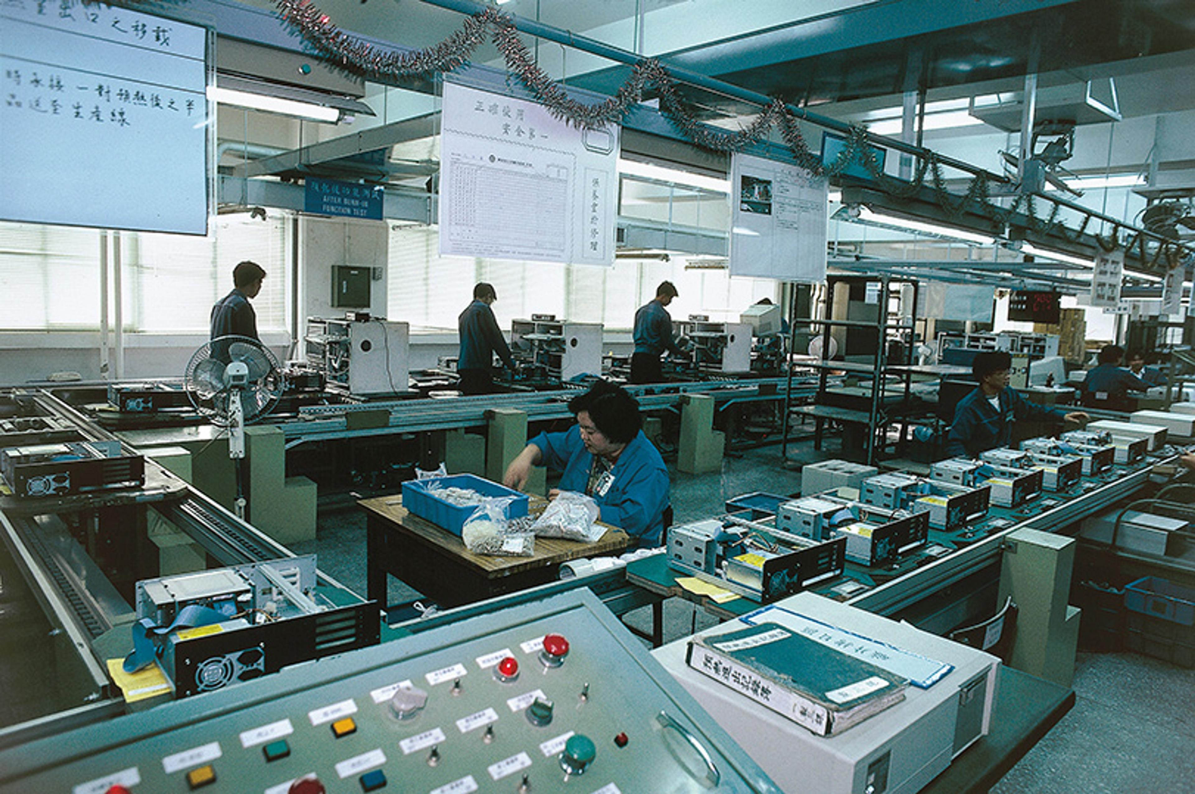 A factory assembly line with workers handling electronics, various equipment and control panels visible.
