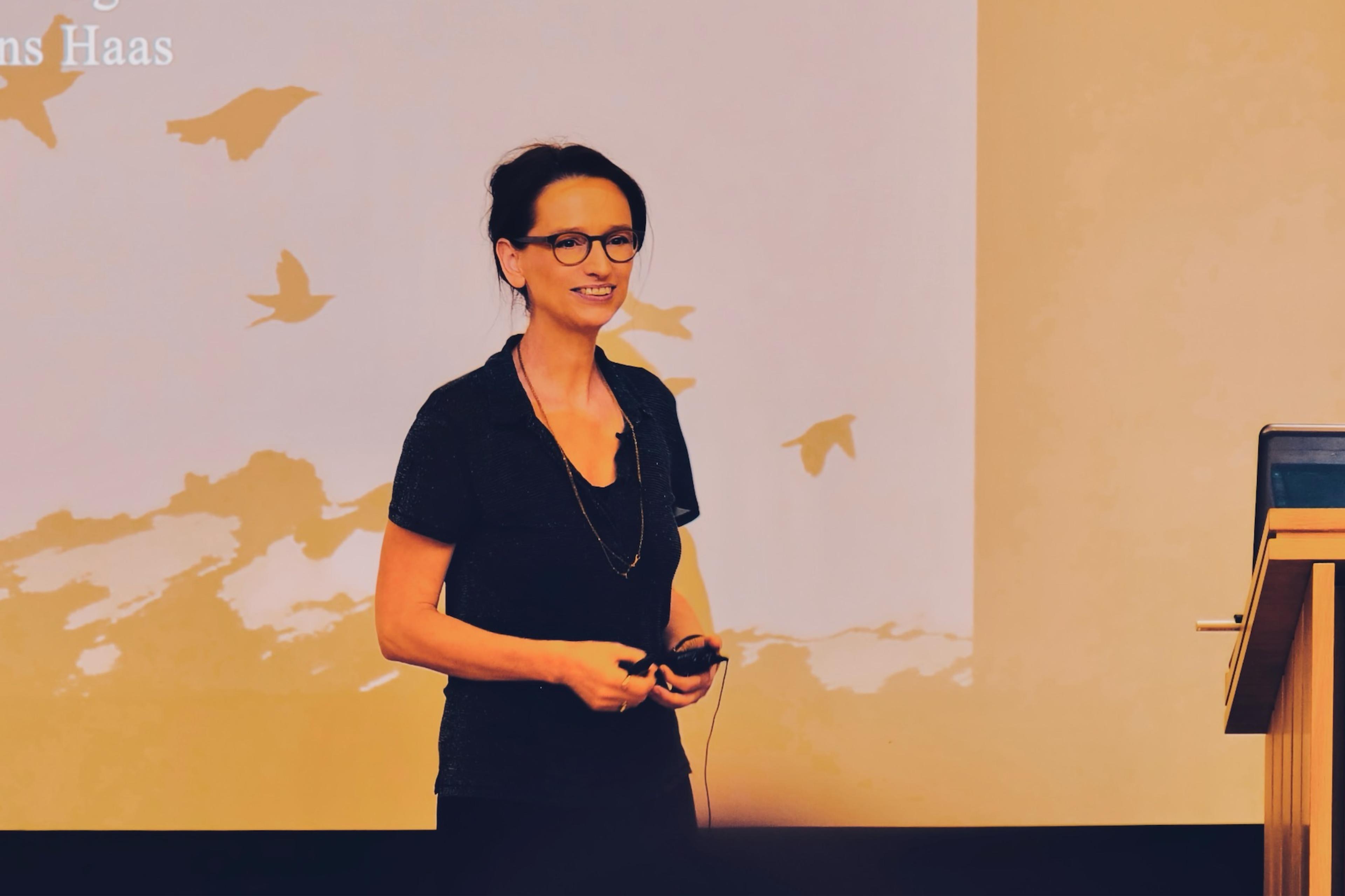 Photo of a woman speaking at a podium with a background showing silhouettes of birds in flight.