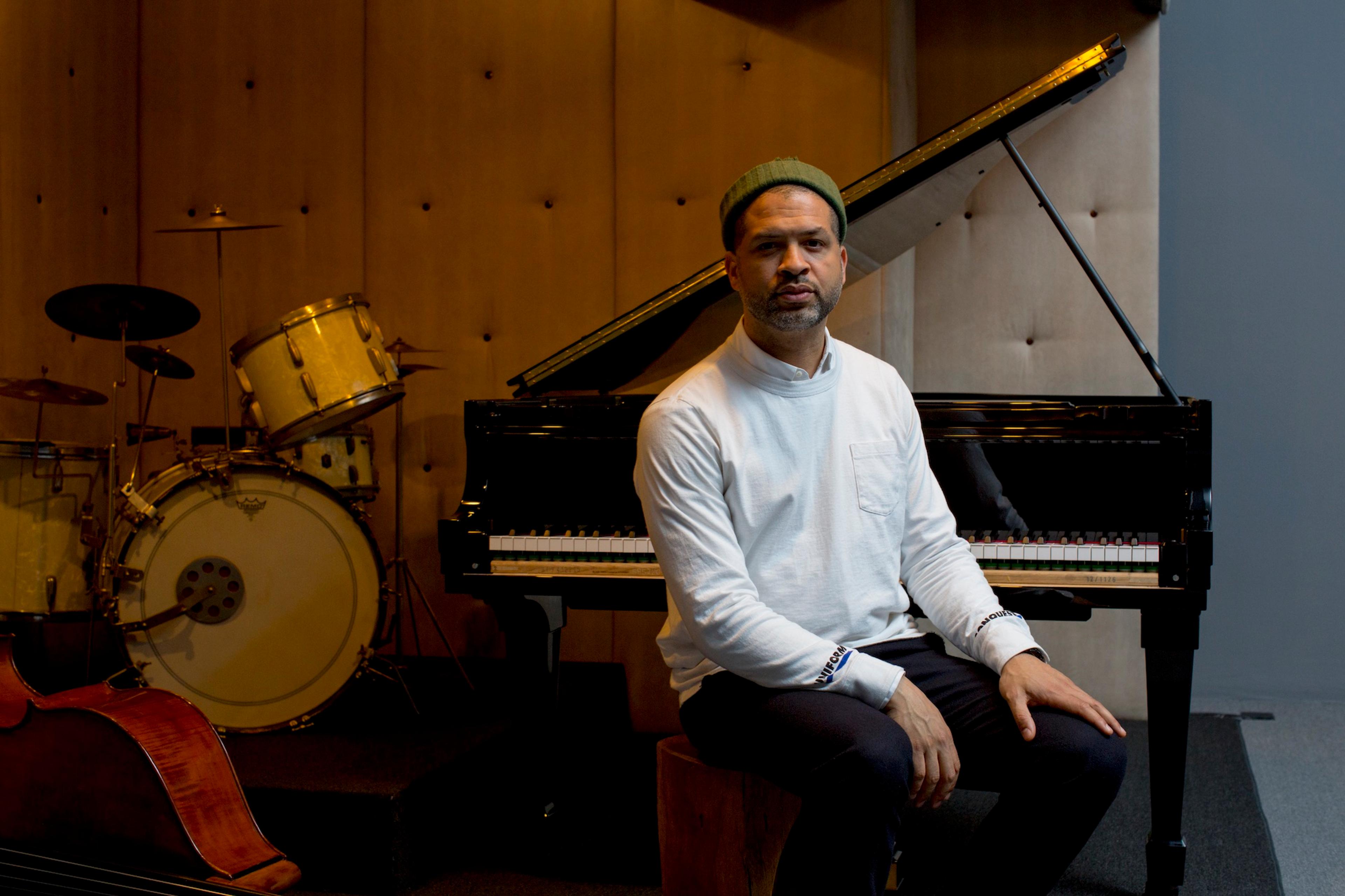 Photo of a person seated by a grand piano with a drum set and double bass in the background in a music studio.