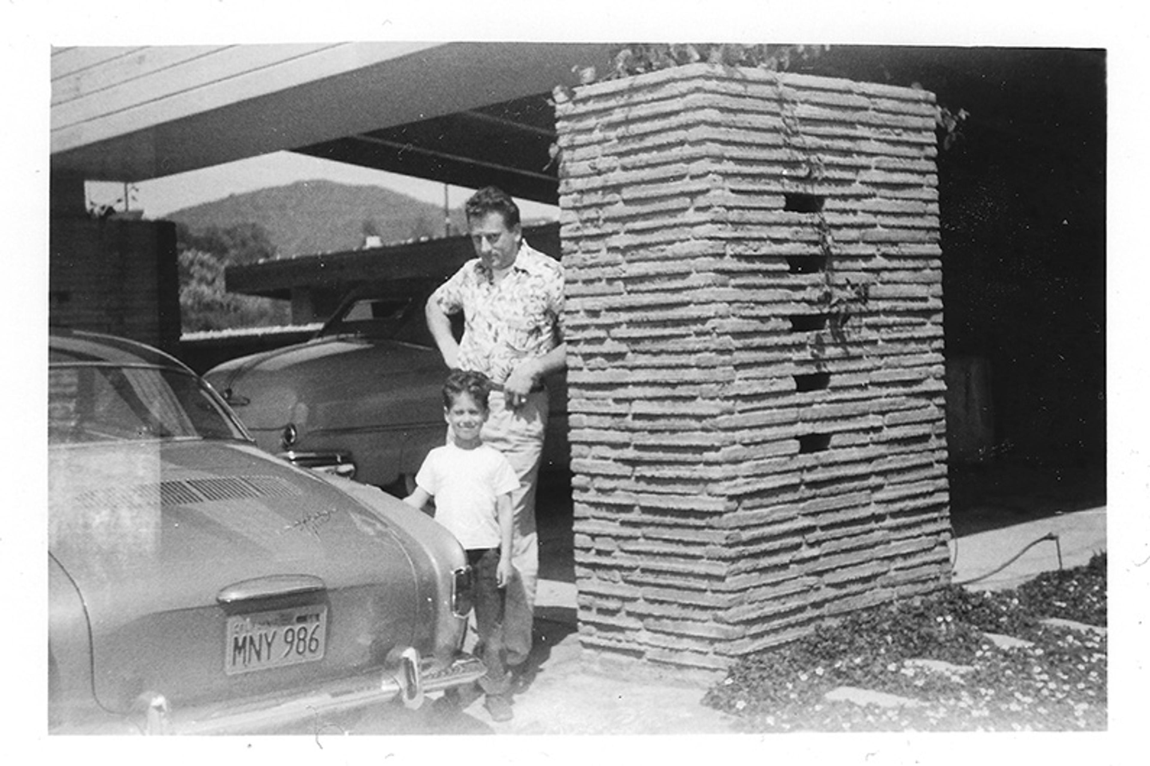 Black and white photo of a man and child near a car by a brick pillar under a carport, mountains visible in background.