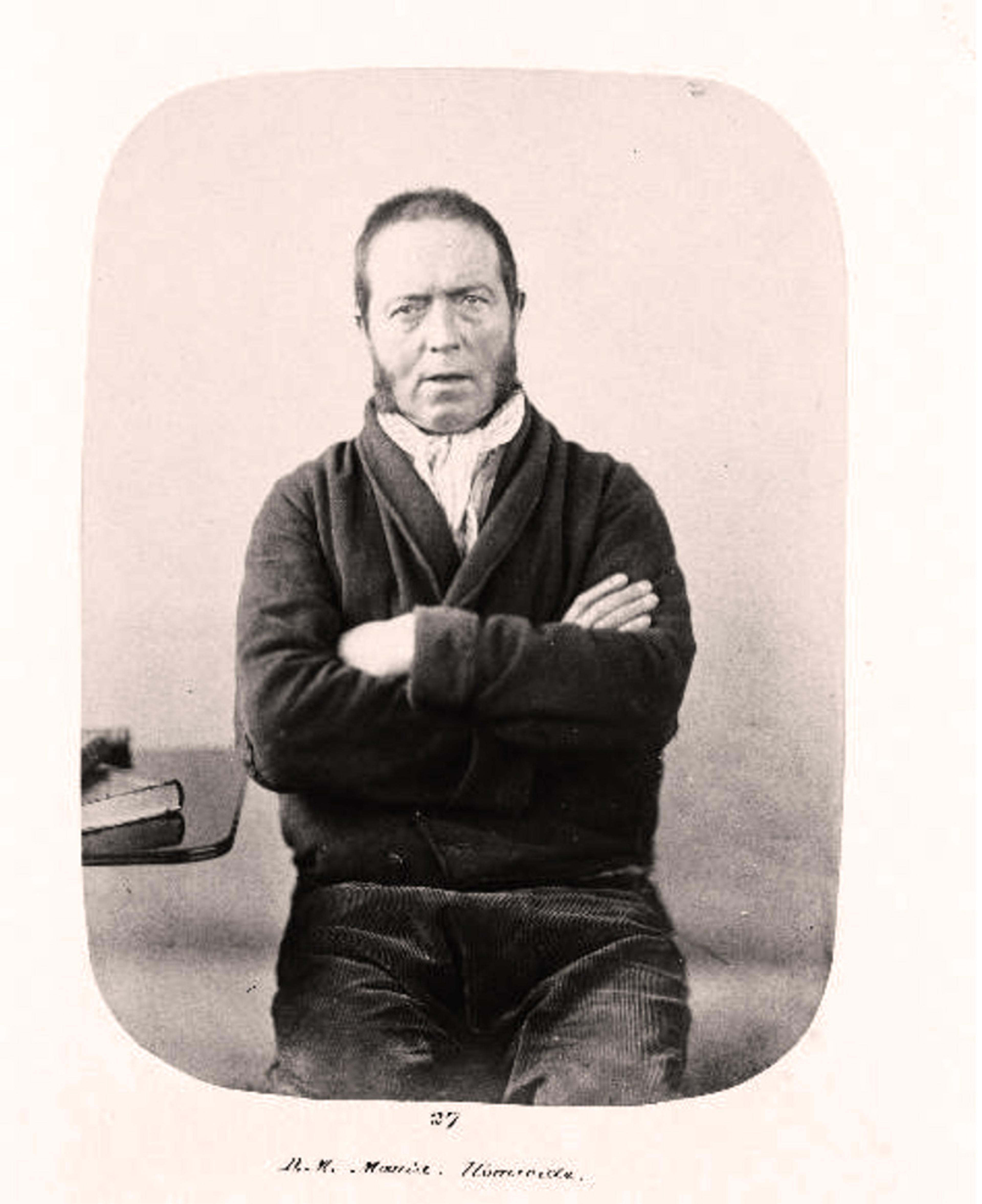 19th-century black and white photo of a man sitting with arms crossed wearing a jacket next to a table with a book.