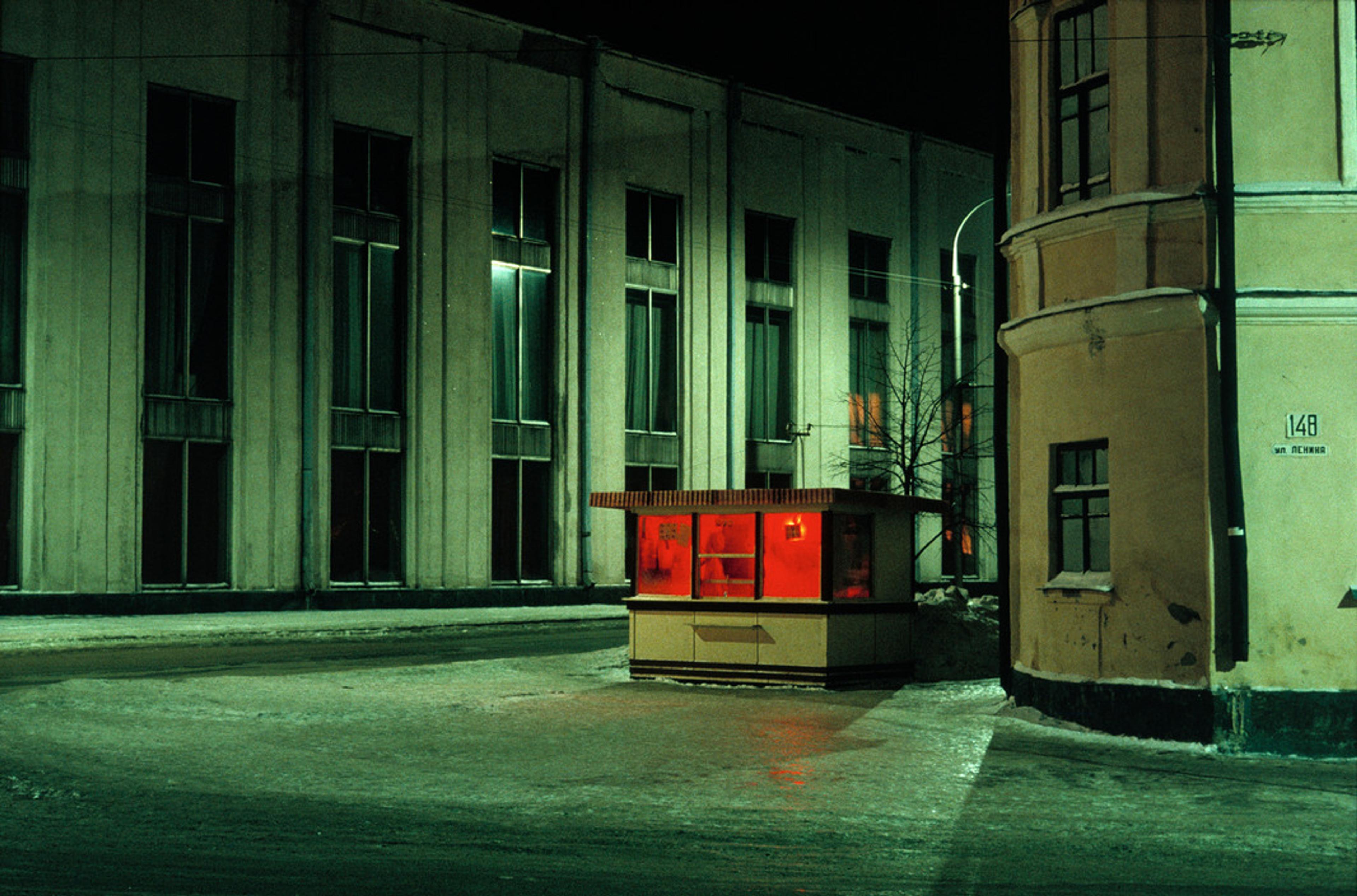 A dimly lit urban street at night with a small building glowing with red light, surrounded by larger buildings.