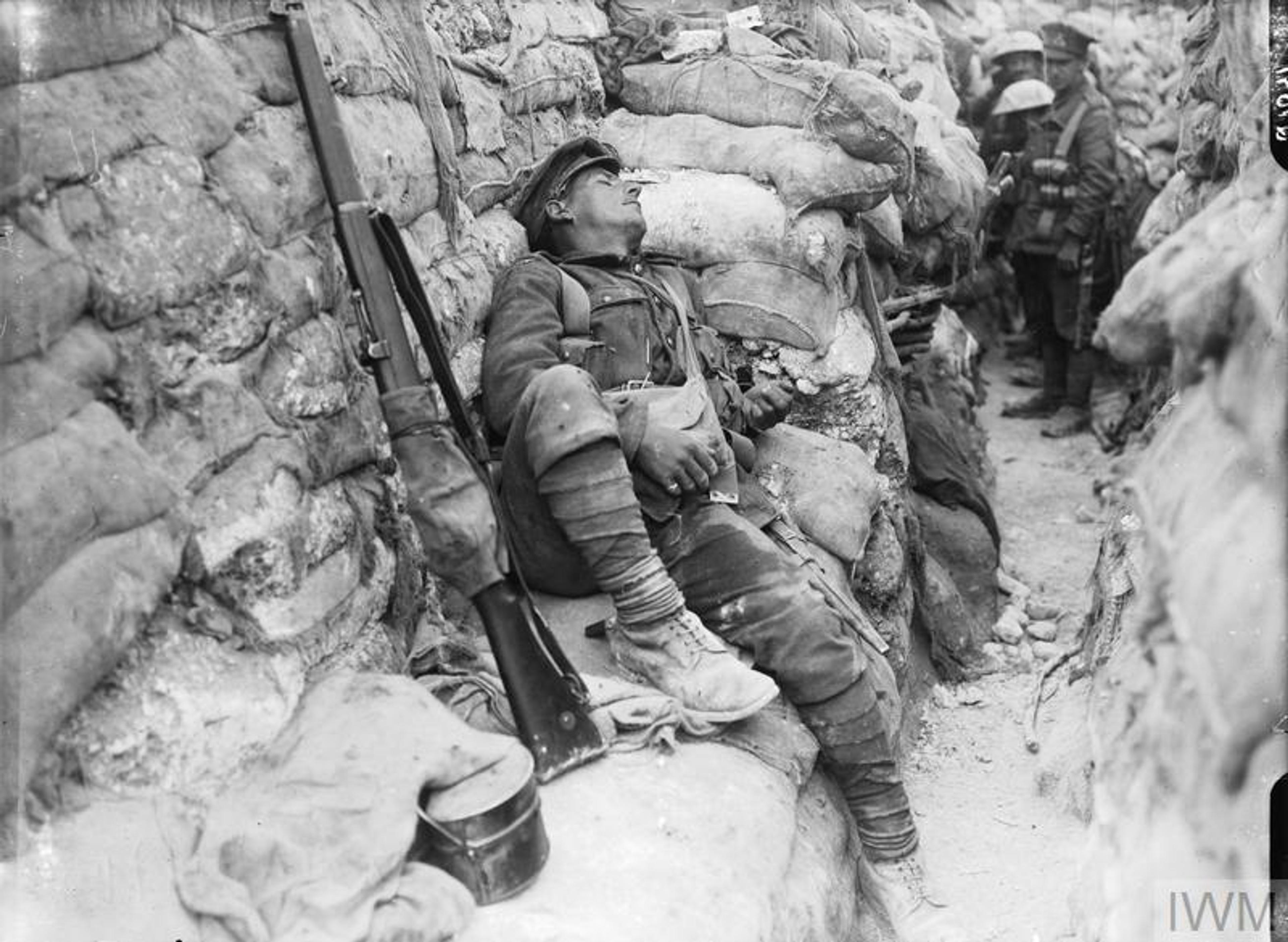 Black and white photo taken during the First World War of a soldier asleep in a trench with a rifle resting nearby and other soldiers visible in the background.