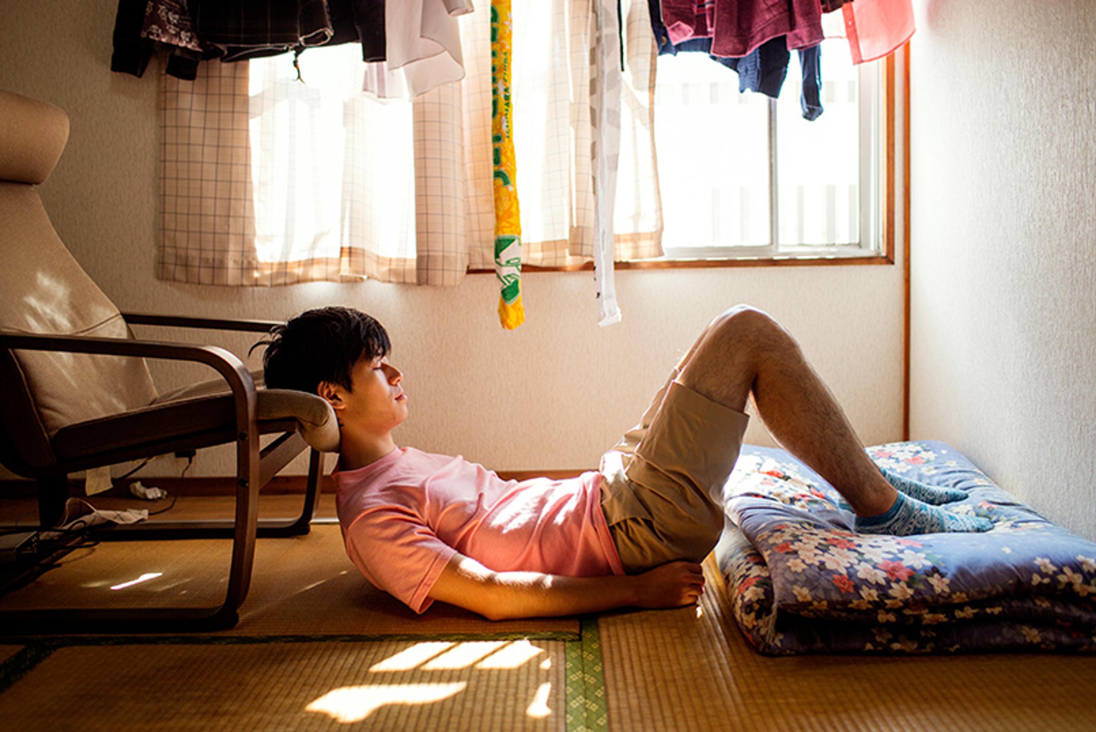 Photo of a person in a pink shirt relaxing on the floor with a cushion in a sunlit room with hanging clothes.